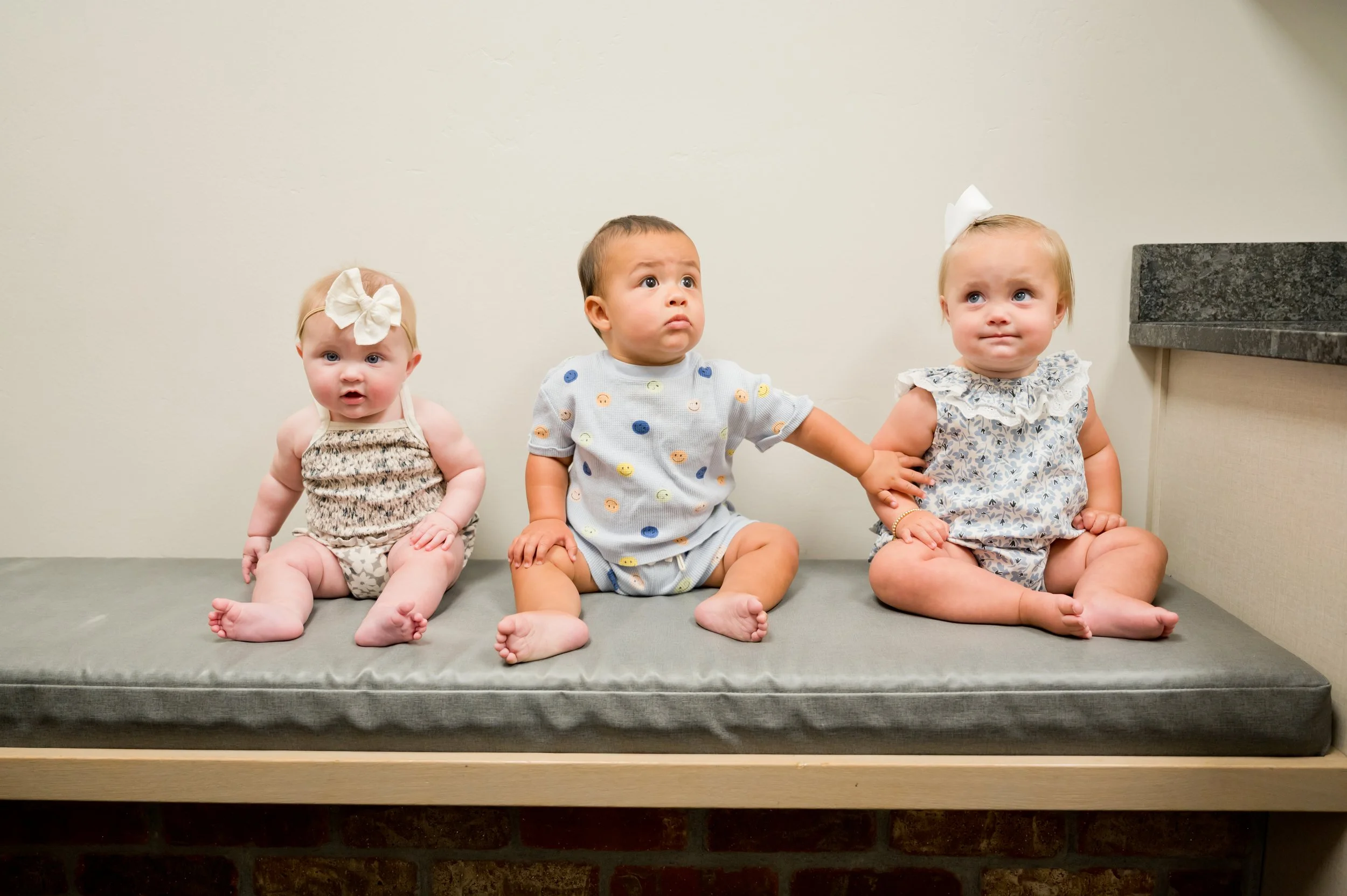 Three babies are sitting on a padded bench against a plain wall, with the middle baby reaching out and touching the baby on the right.