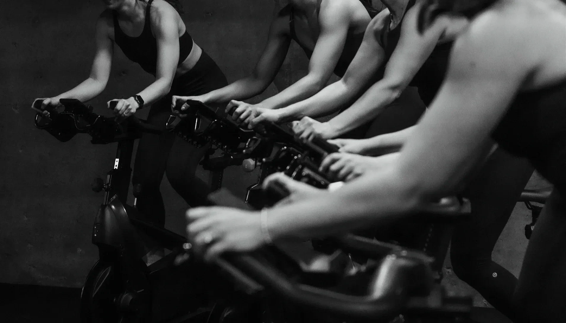 Group of women in athletic clothing participating in a spin class, riding stationary bicycles in a dark indoor setting.