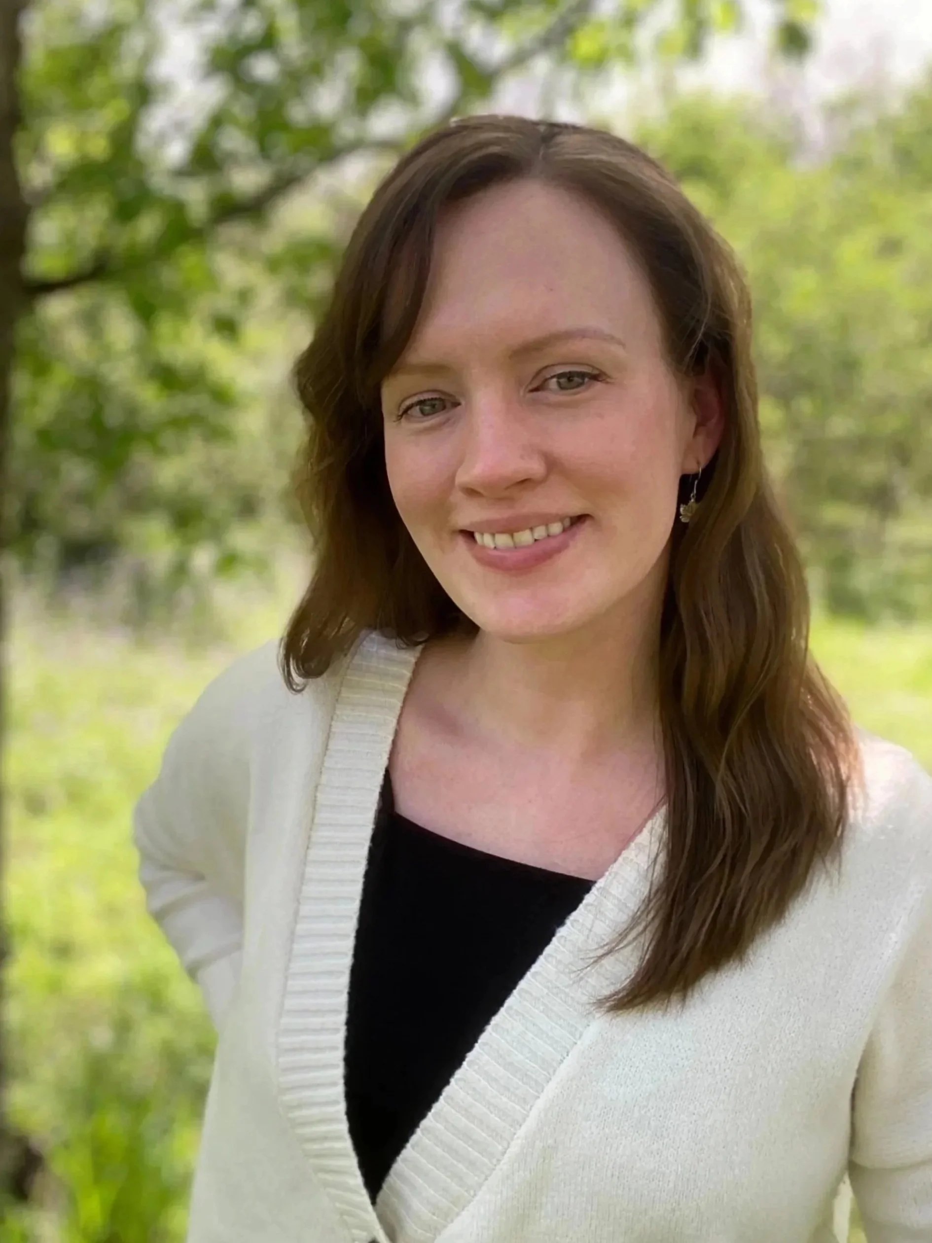 A woman with long brown hair smiling outdoors, wearing a cream cardigan over a black top, with a background of green trees and sunlight.