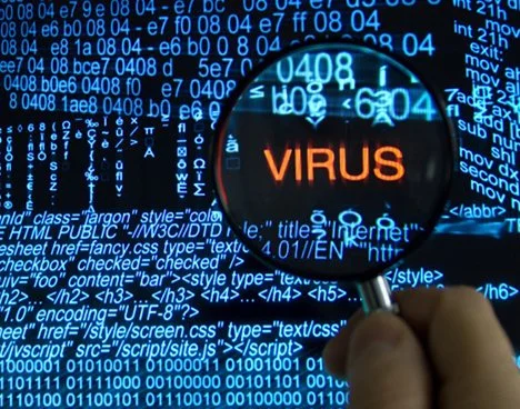 A person's hand holding a magnifying glass over a computer screen displaying digital code and the word 'VIRUS' highlighted, symbolizing cybersecurity or malware analysis.