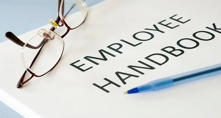 A pair of reading glasses, a blue pen, and a printed employment handbook on a light-colored surface.