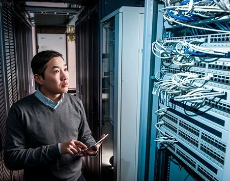 A woman standing in a data center, holding a tablet, with server racks and networking equipment around her.