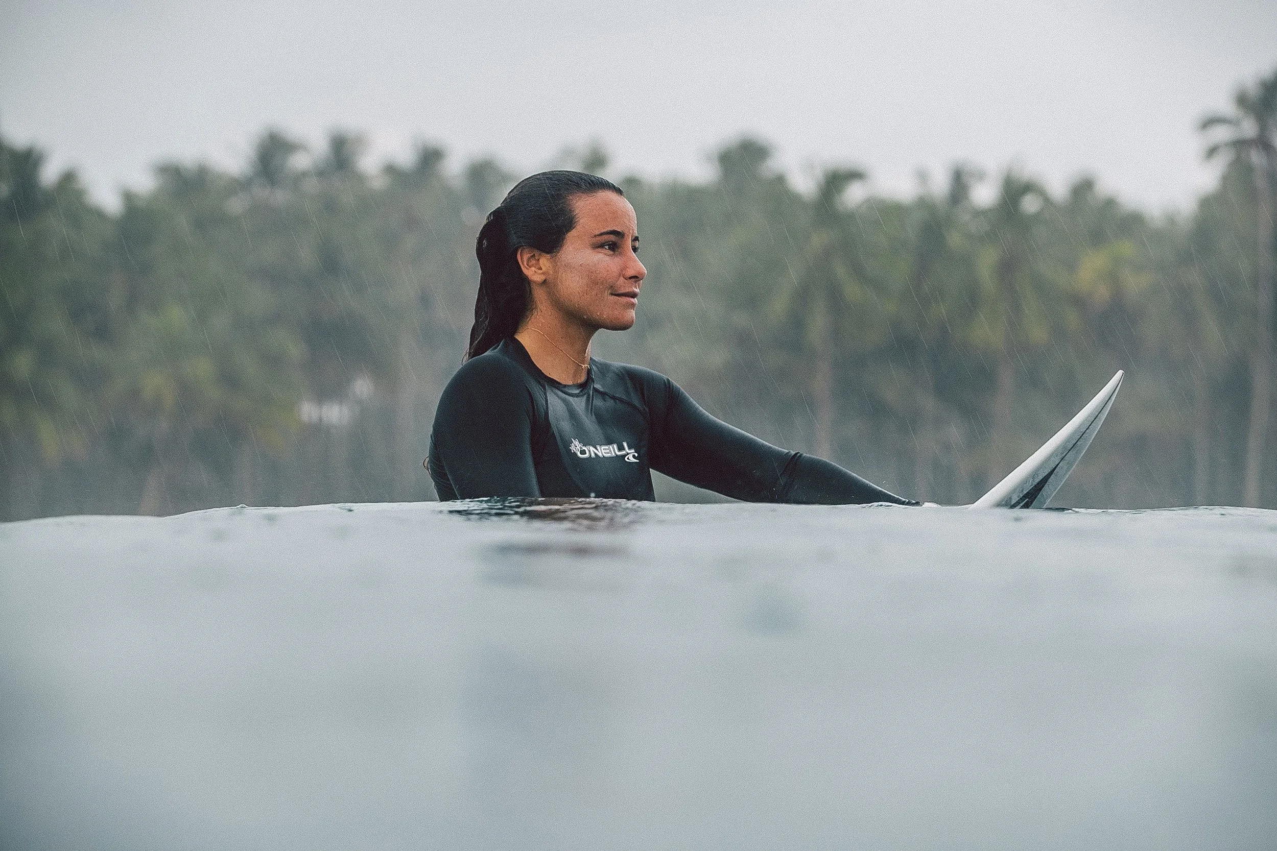 A woman in a wetsuit sitting on a surfboard in the water, with rain falling and a background of trees.