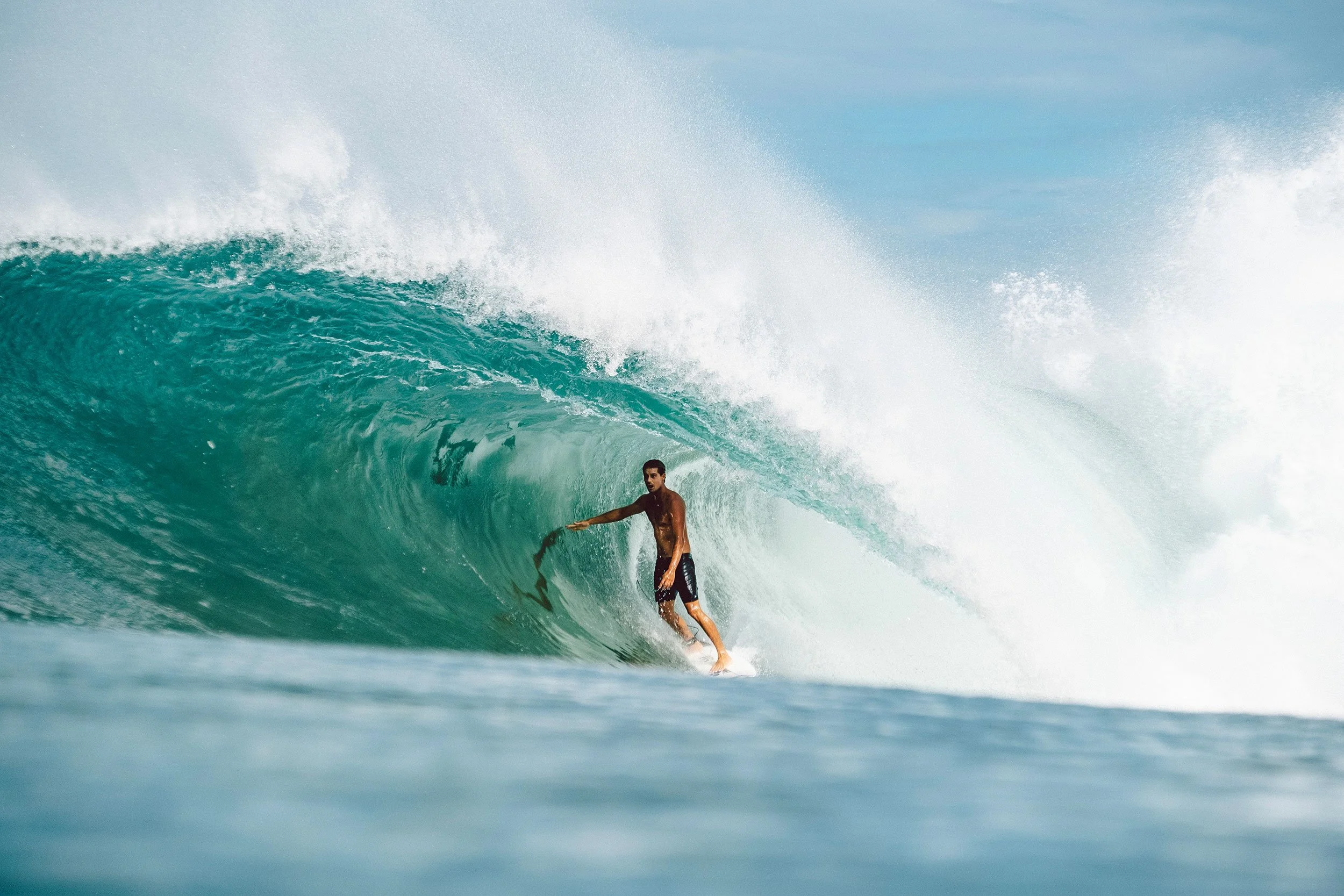 A man surfing inside a large ocean wave with clear blue water and a partly cloudy sky.