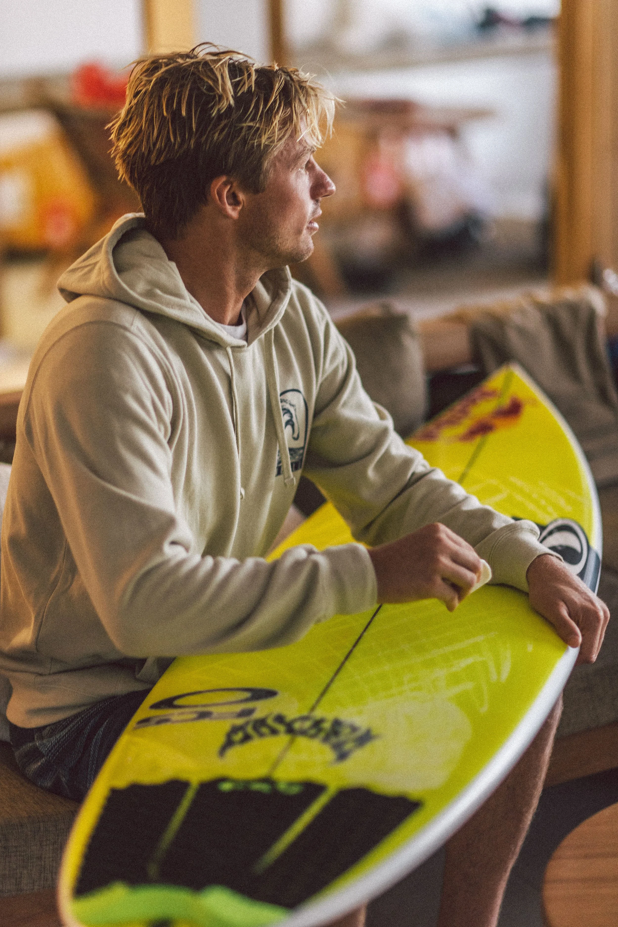 A young man with blond hair, wearing a beige hoodie, sitting indoors and holding a yellow surfboard.