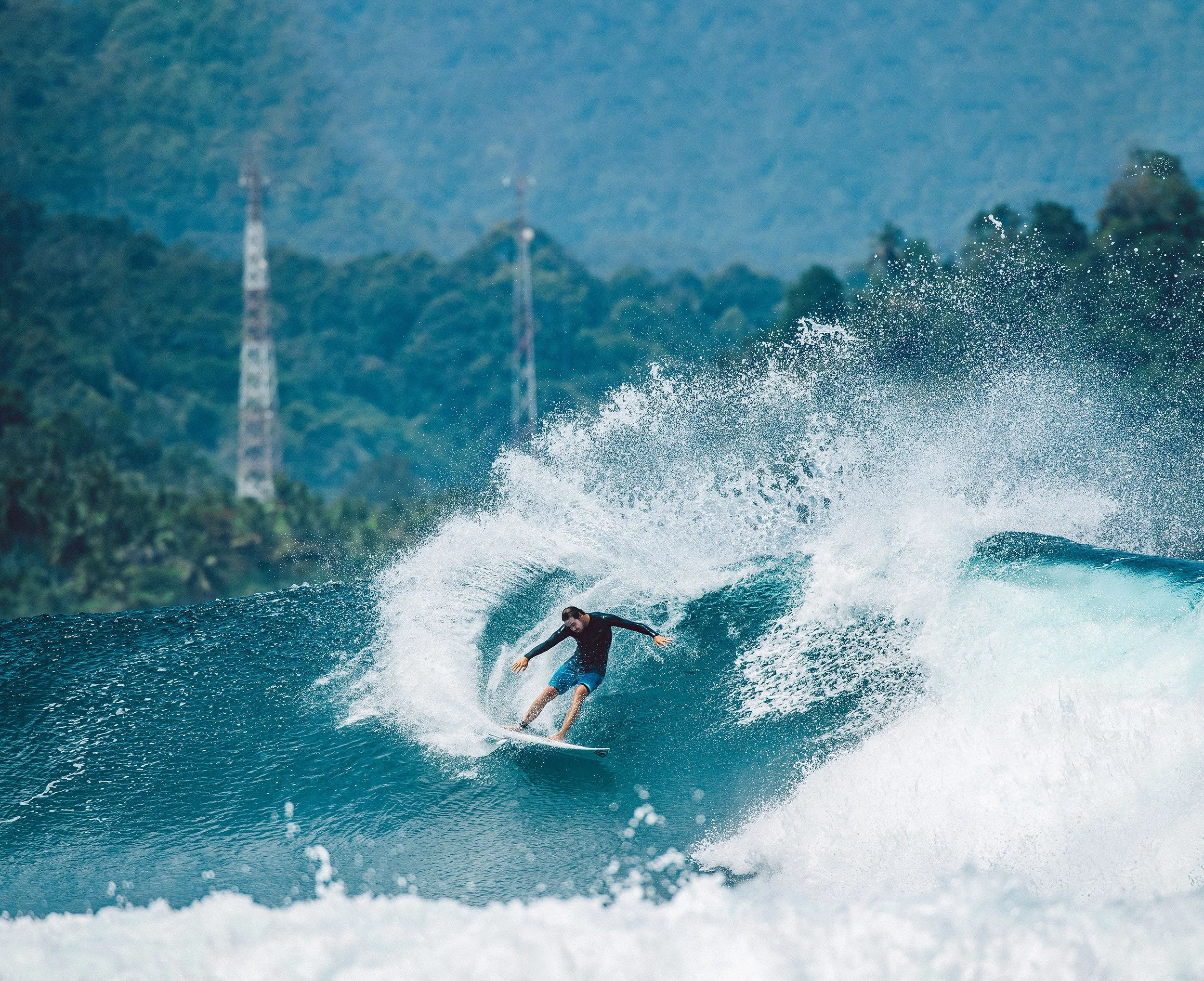 A person surfing on a wave in the ocean with a forested hillside and communication towers in the background.