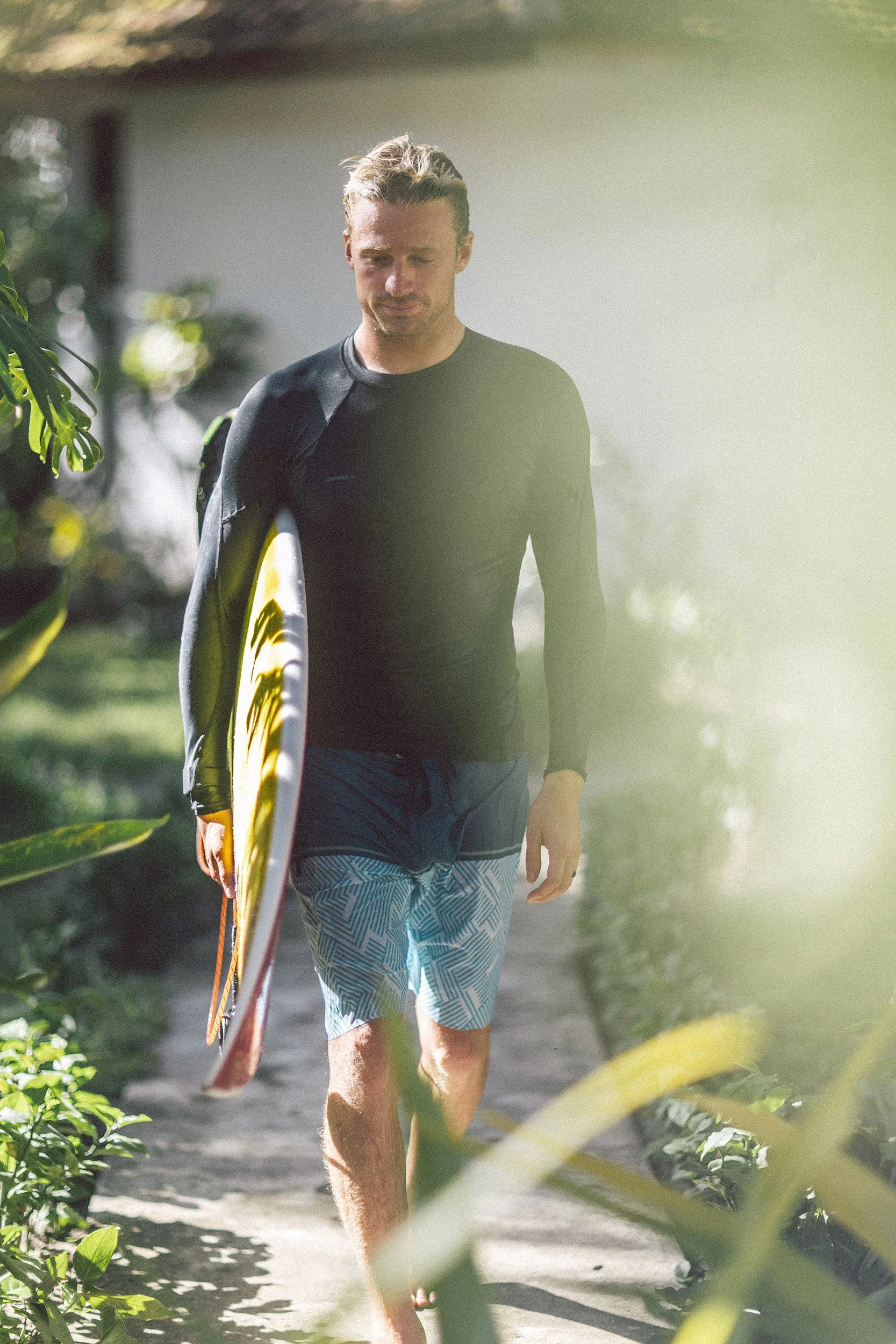 A man walking along a sandy path carrying a surfboard under his arm, dressed in a long-sleeve rash guard and patterned board shorts.