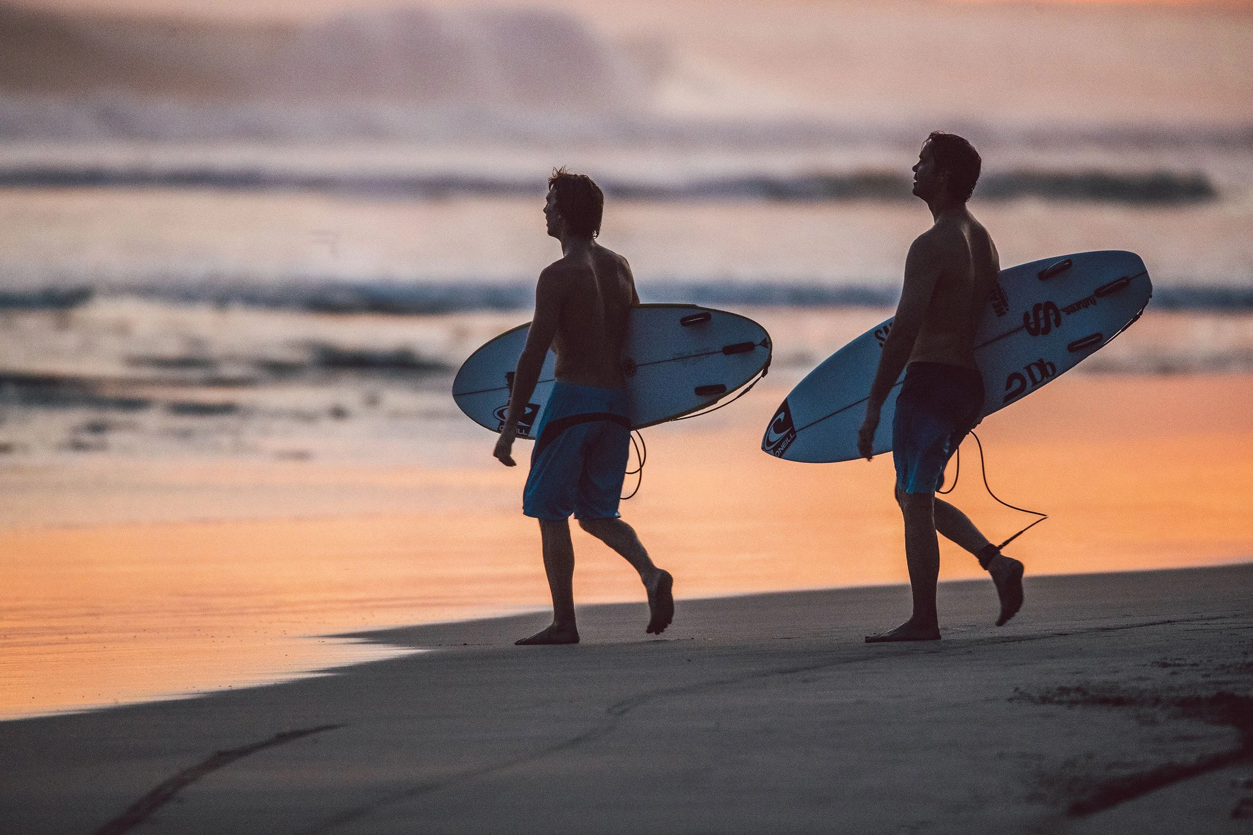 Two men carrying surfboards walk along the beach at sunset, with the ocean waves in the background.