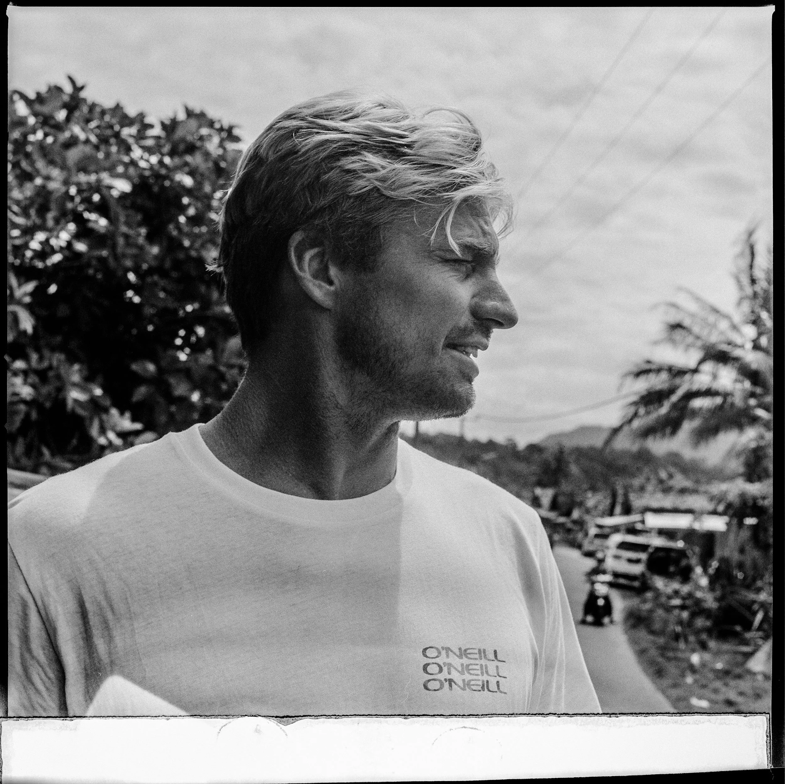 Black and white photo of a man with blonde hair and a beard, wearing a t-shirt, standing outdoors with trees and cars in the background.