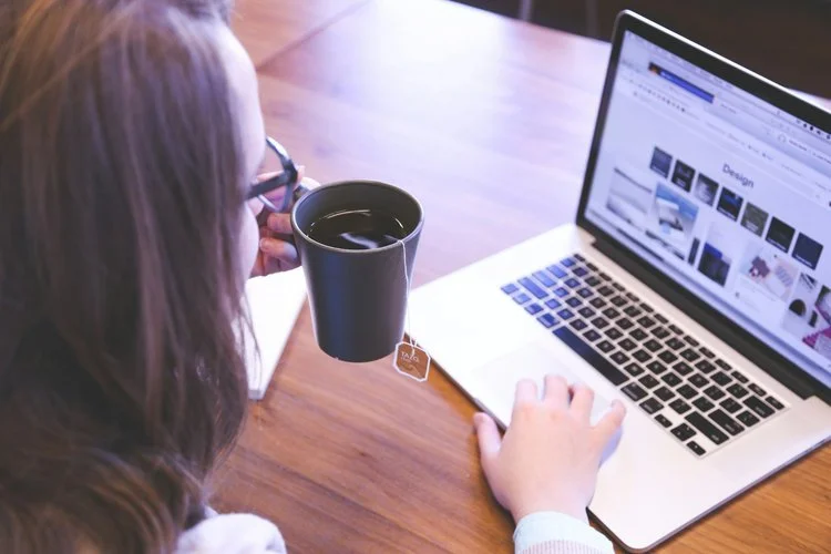Woman holding a cup of tea and working on a laptop.