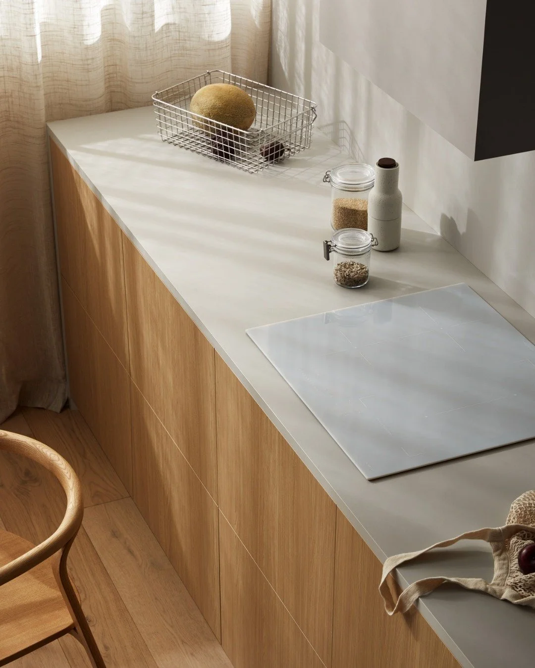 Kitchen countertop with a wire basket containing a cantaloupe, jars of grains and oats, a spice container, and a white cooktop.