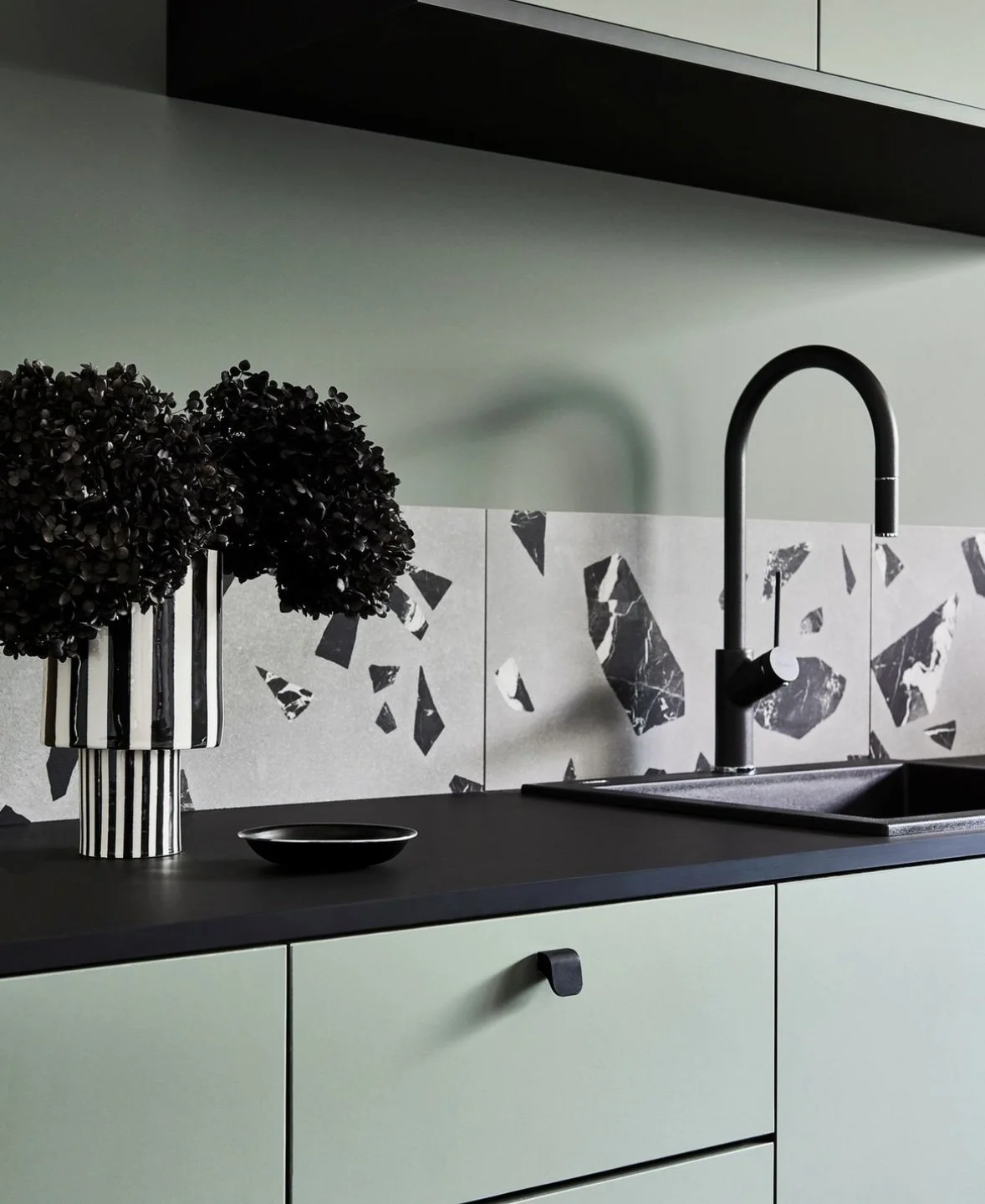 Modern kitchen countertop with black sink, black faucet, leafy black plant in striped vase, small black dish, and patterned tile backsplash with black and white abstract shards.