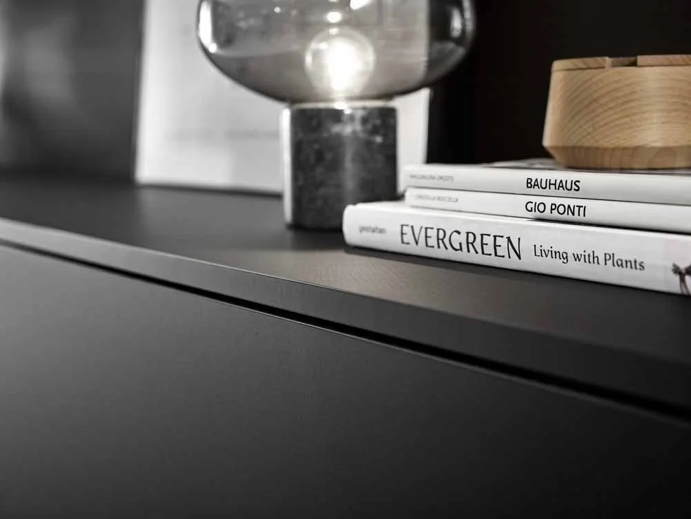 Close-up of a black bookshelf or cabinet top with three books, a glass vase, and a wooden bowl, against a blurred background.