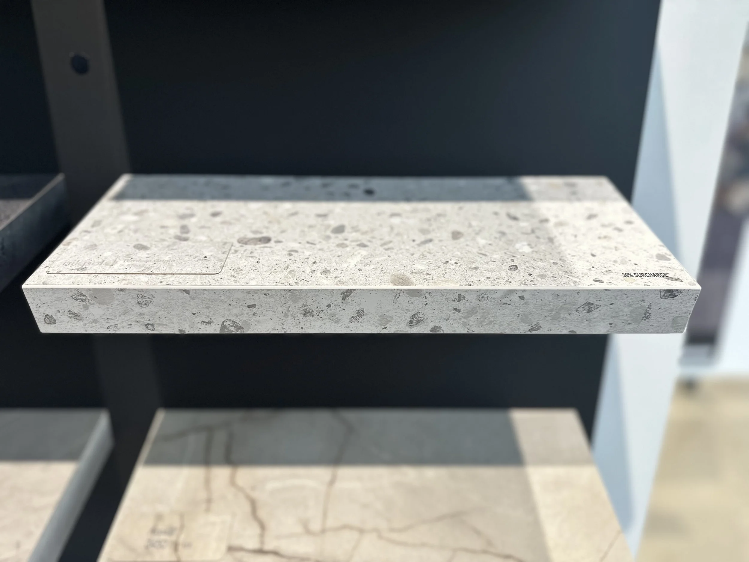 A display board showcasing laminate profiles with a speckled pattern, mounted on a black wall. The shelf has a small label on the right side and a square cover on the left side. The background shows a blurred interior.