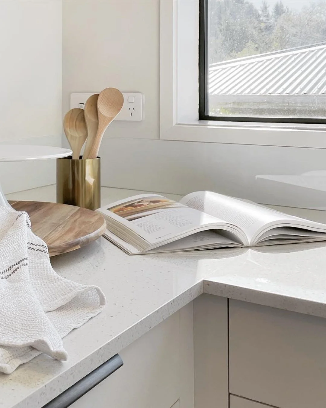 Open cookbook on white kitchen counter with wooden cutting board, white dish towel, and a gold container holding wooden spoons near a window.