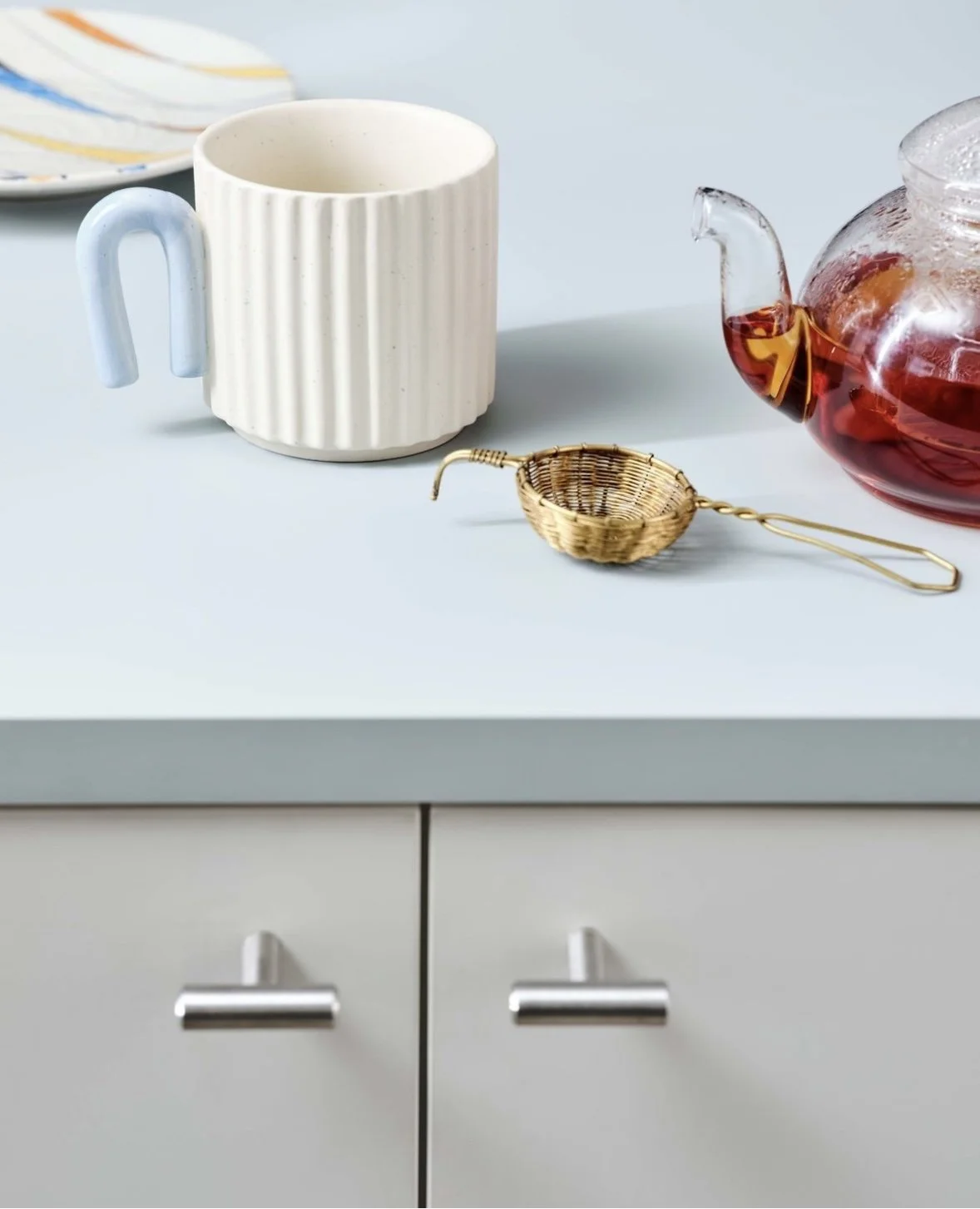A white mug, a glass teapot with red tea, a gold coffee filter, and a white kitchen drawer with silver handles.