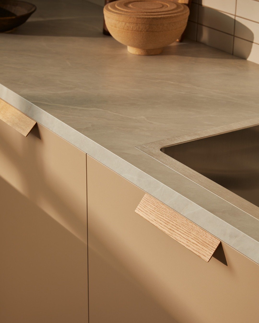 Close-up of a modern Laminex kitchen counter with a beige cabinet, a light countertop, and a built-in sink. Decorative bowls are on the counter, and the background features a tiled wall.