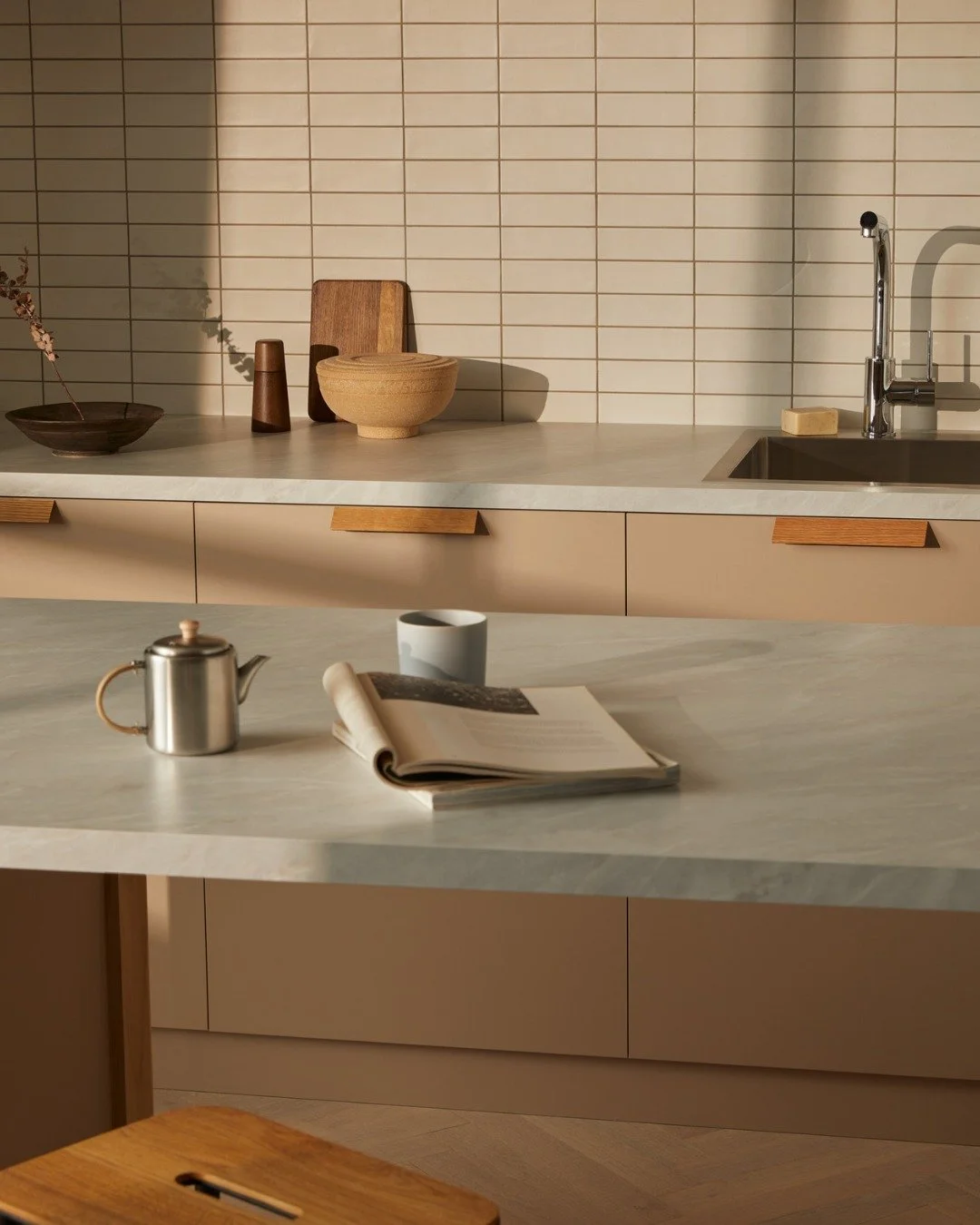 A modern kitchen with a beige countertop, white tiled backsplash, and minimal wooden decor. Items on the counter include a wooden bowl, a small wooden cutting board, a ceramic bowl, a ceramic mug, a teapot, and a newspaper. The sink has a chrome fauc