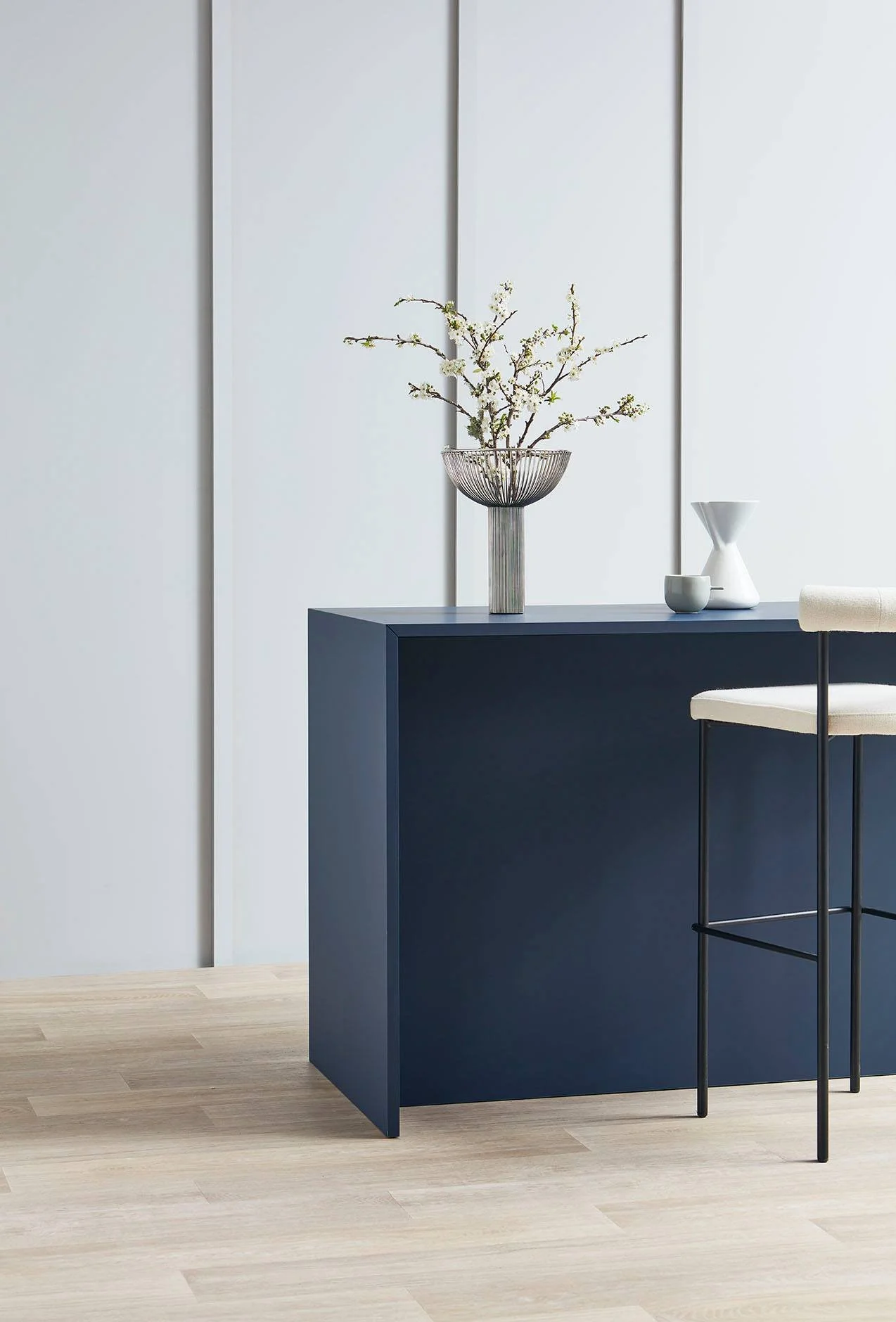 A minimalist interior with a navy blue table, a white, black-legged stool, a metal vase with white flowers, and white ceramic decor on the table against a white wall with vertical paneling.