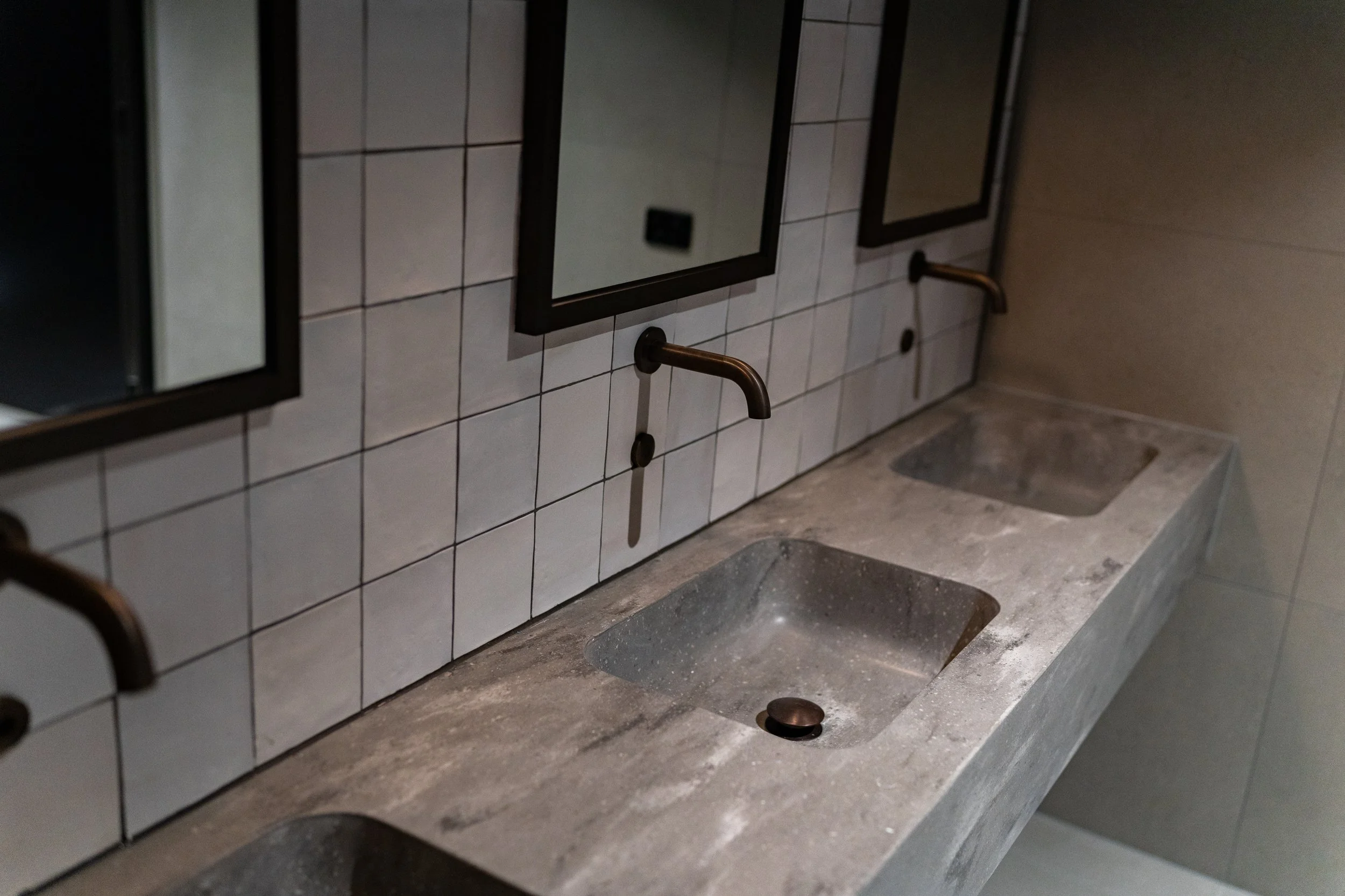 A public restroom with three corian integrated basins into the benchtop, three wall-mounted faucets, three mirrors above the sinks, and tiled walls.