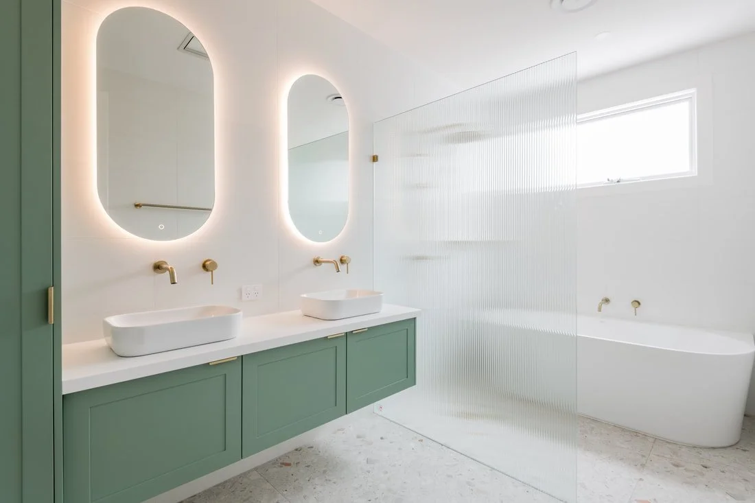 Modern bathroom with corian glacier white benchtop with double vanity, two oval mirrors, green cabinets, brushed brass fixtures, a frosted glass partition, and a freestanding bathtub near a window.