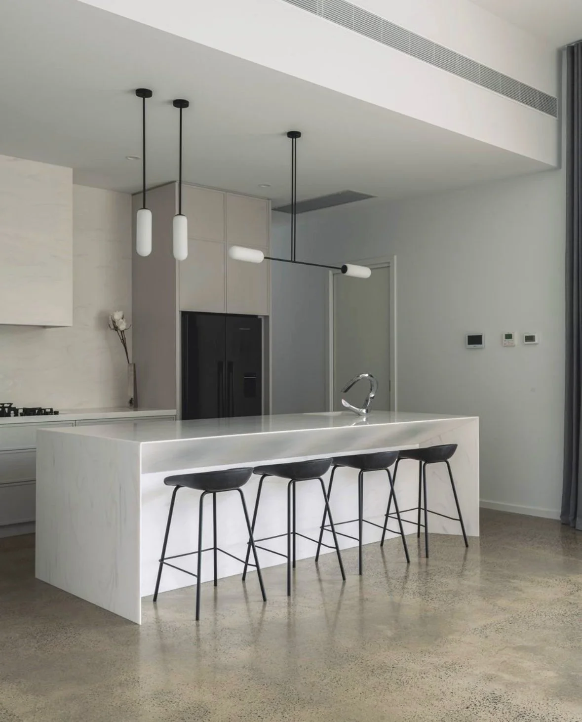 Minimalist kitchen with a corian venaro white island, three black stools, modern pendant lighting, and a black refrigerator amidst neutral tones.