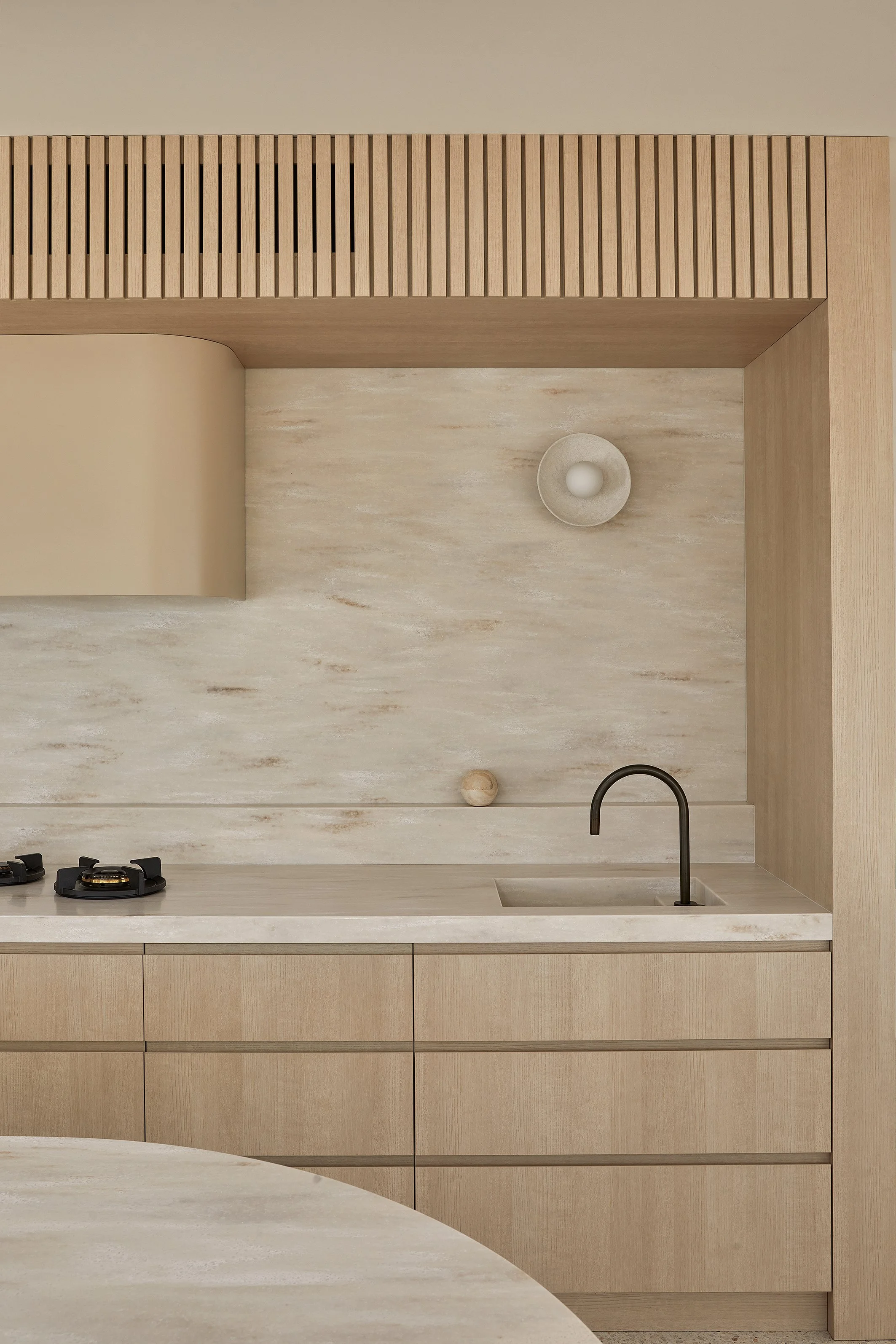 A minimalist kitchen with timber cabinets, corian witch hazel benchtops and splashback, a small sink with a gunmetal faucet, a stove with two burners, a wooden backsplash, and a wooden slatted area above.
