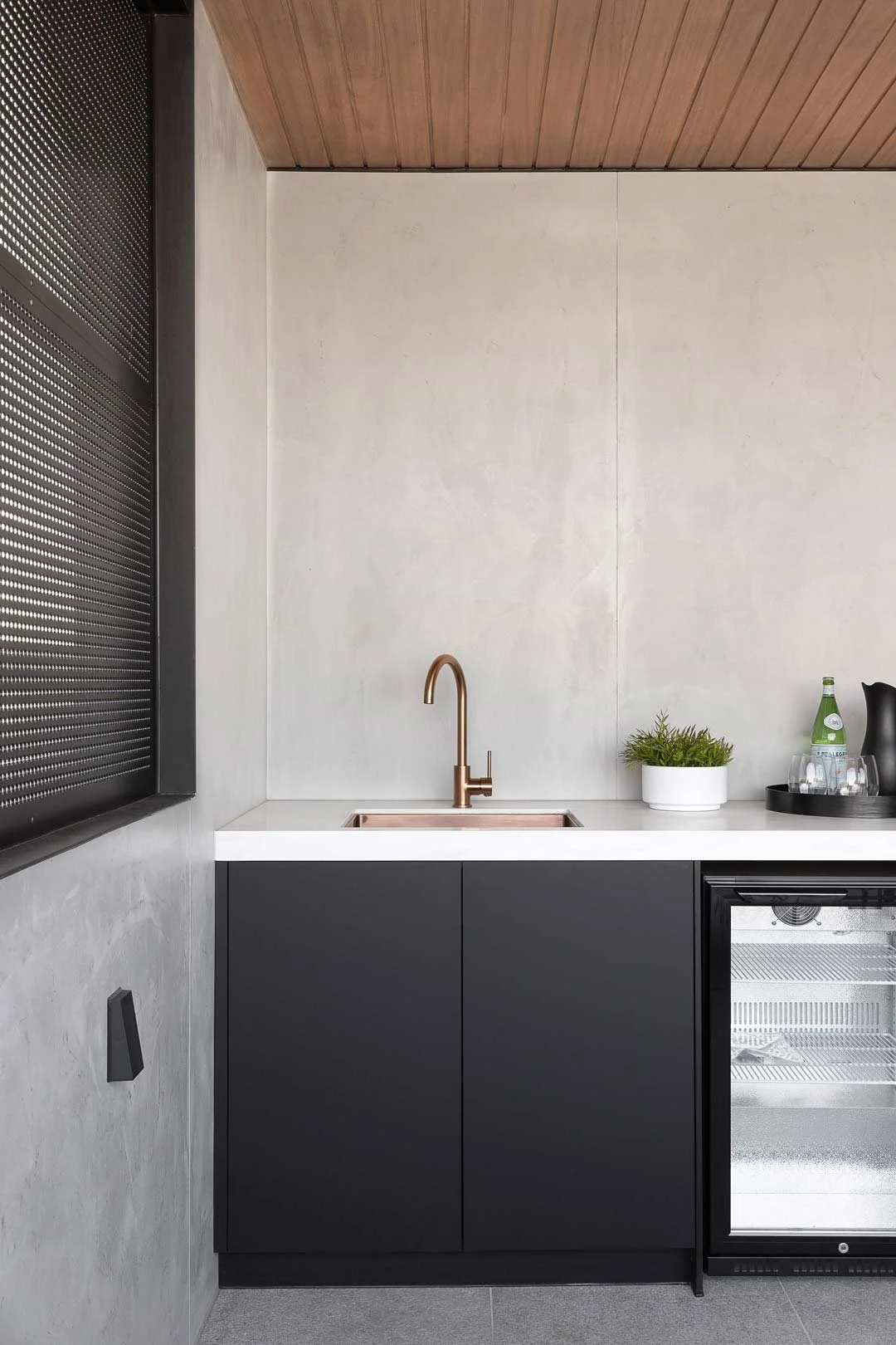 Modern kitchen corner with black cabinets, white countertop, copper faucet, small white plant pot, glass bottle, and a wine fridge.
