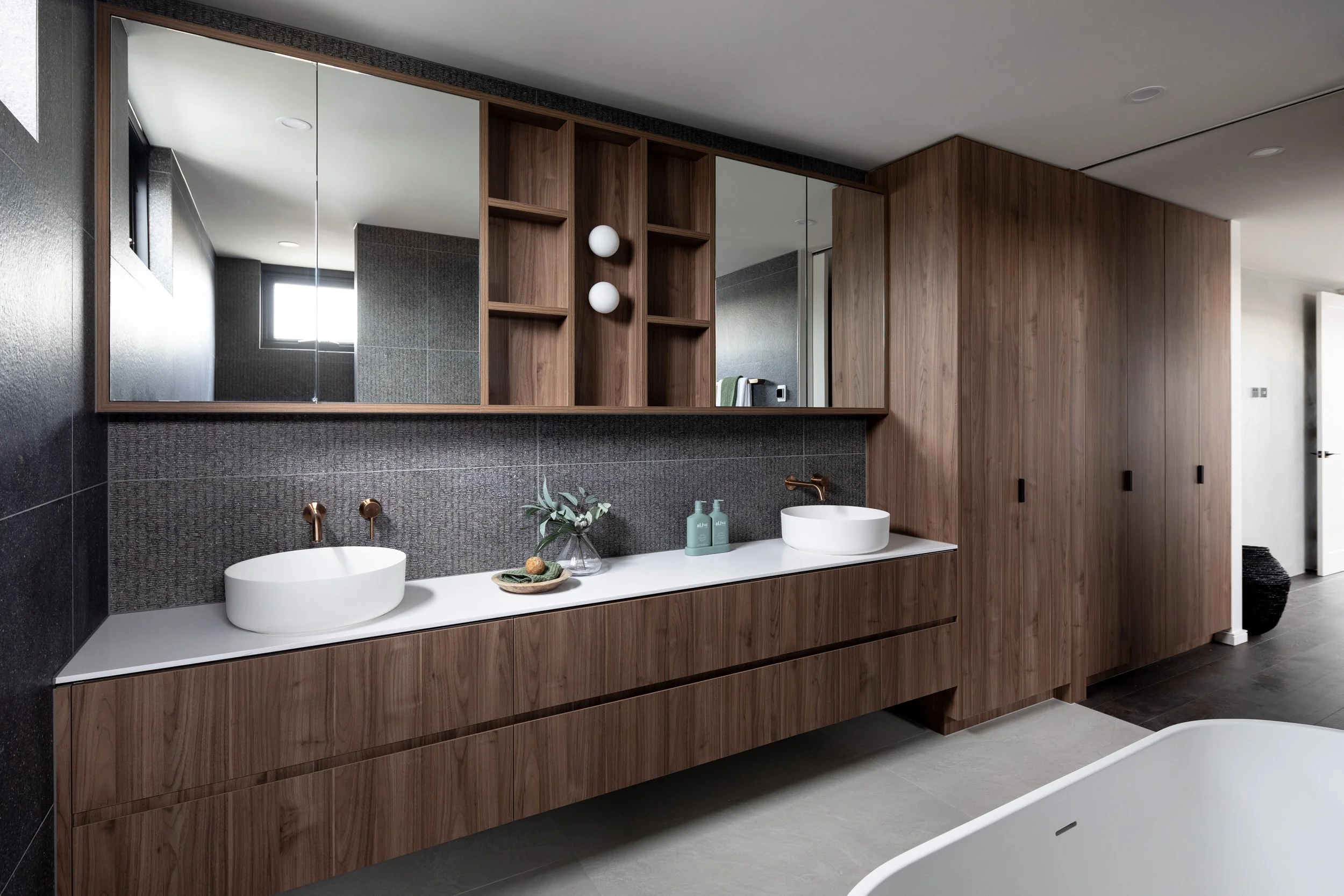 Modern bathroom with double vanity, wooden cabinets, gray textured backsplash, round white vessel sinks, copper faucets, wall-mounted lighting, mirror cabinet, and a freestanding bathtub.