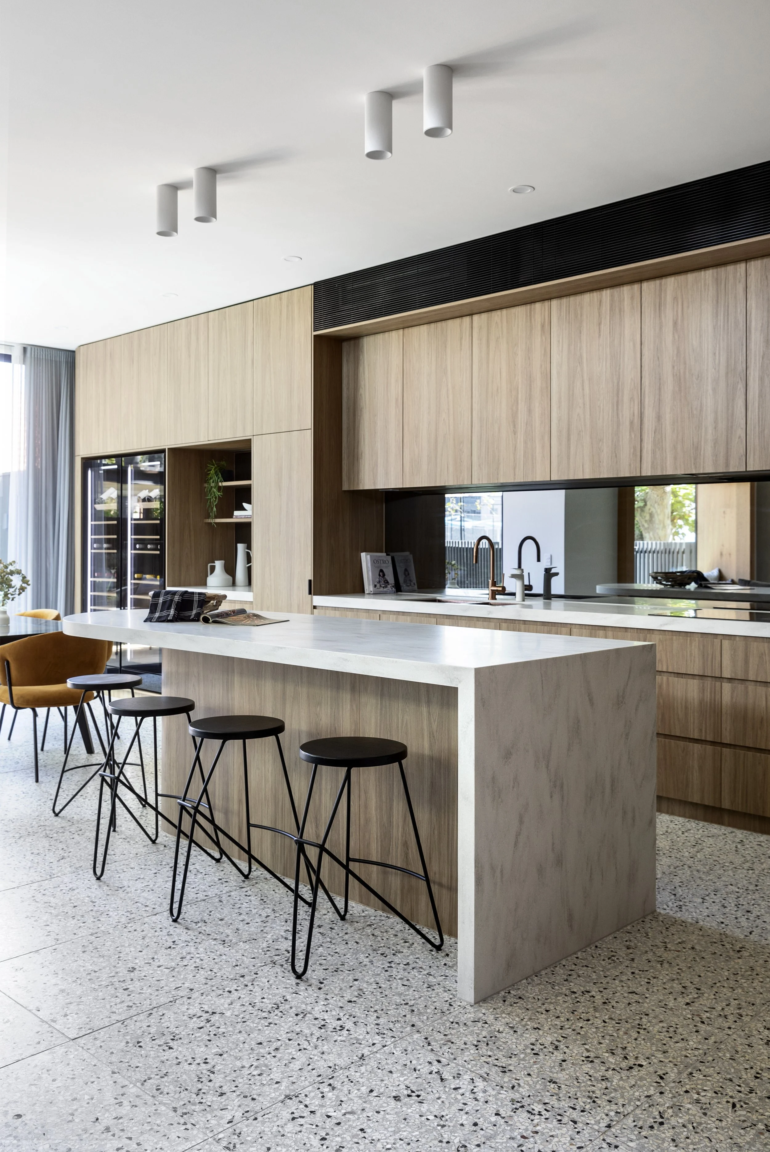 Modern kitchen with wooden cabinets, a corian raincloud island, and black bar stools. Large windows with sheer curtains allow natural light, and a built-in wine fridge is visible. The space features a minimalist design with neutral tones.