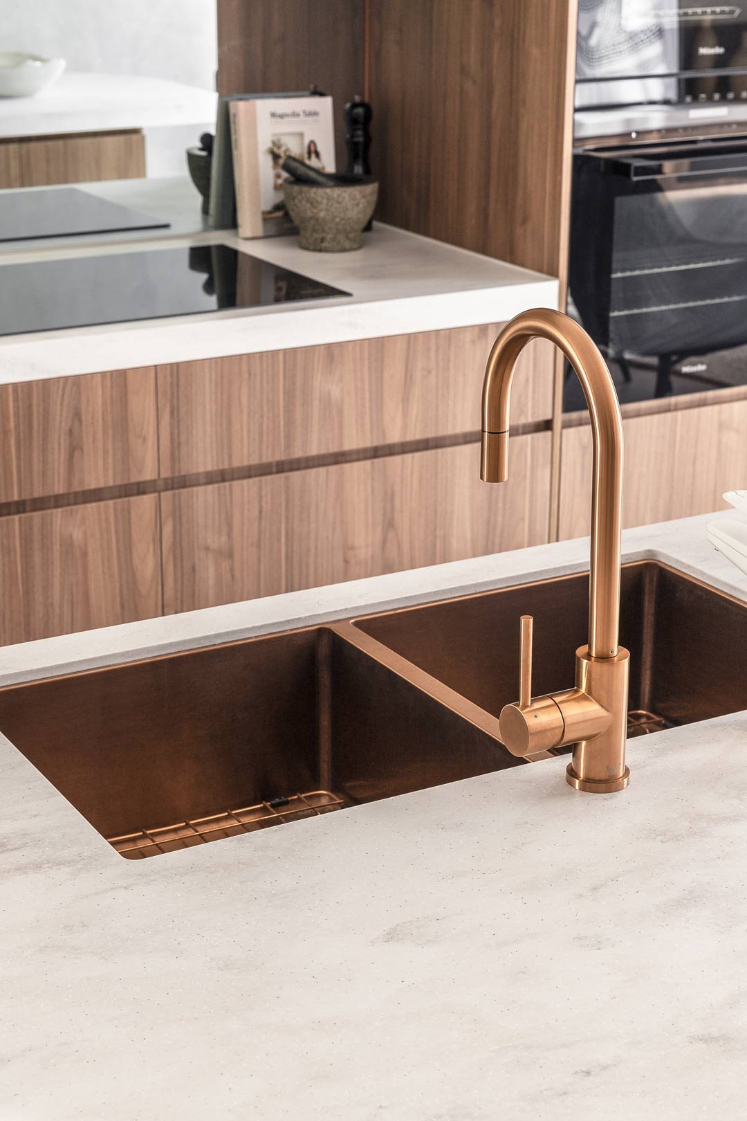 A modern kitchen sink with a copper faucet, set in a light-colored corian raincloud countertop, with a wooden cabinet and stove in the background.