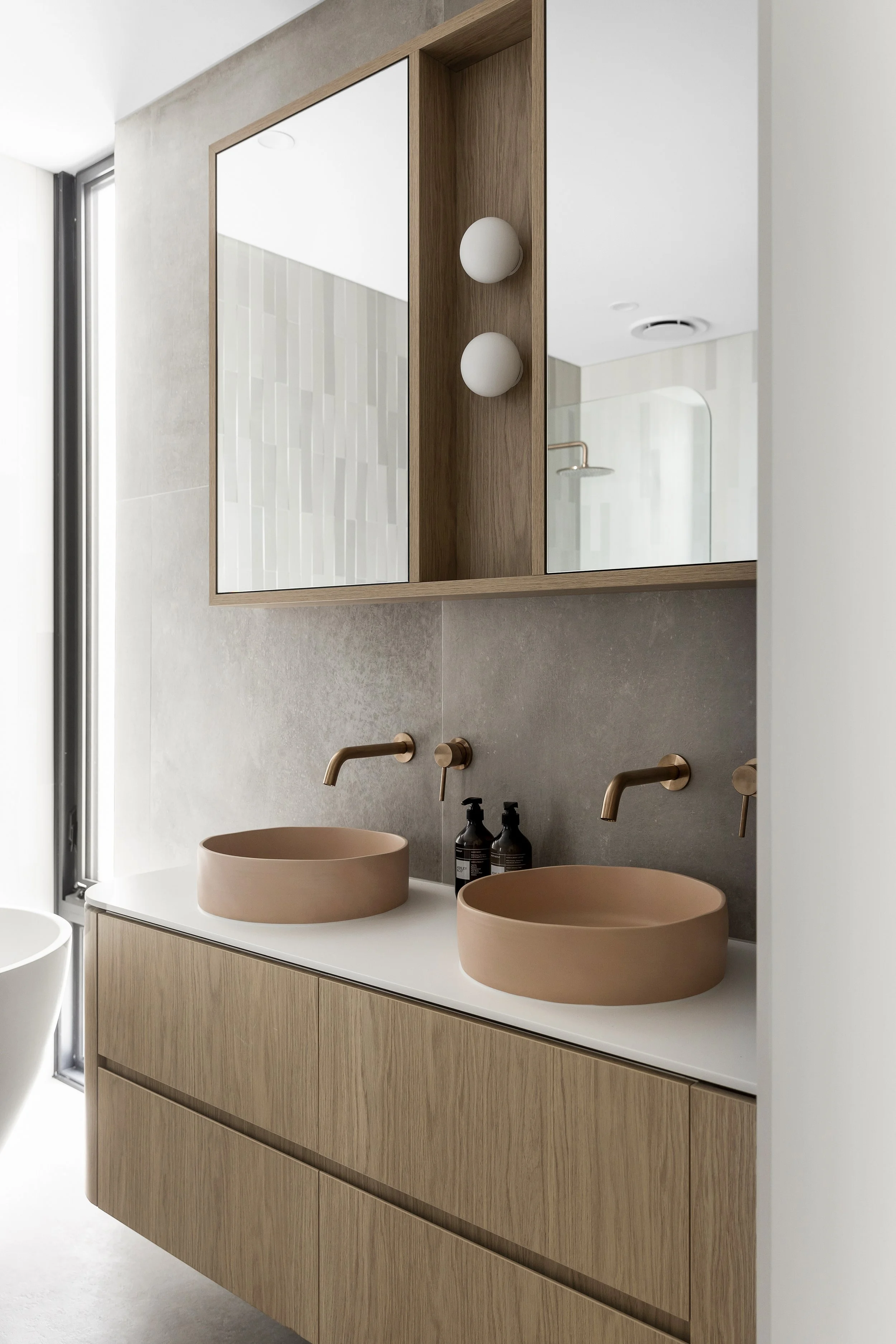 Modern bathroom with corian glacier white vanity benchtop with dual sinks, round vessel basins in a neutral beige color, wall-mounted faucets, a large mirror, and wooden cabinetry.