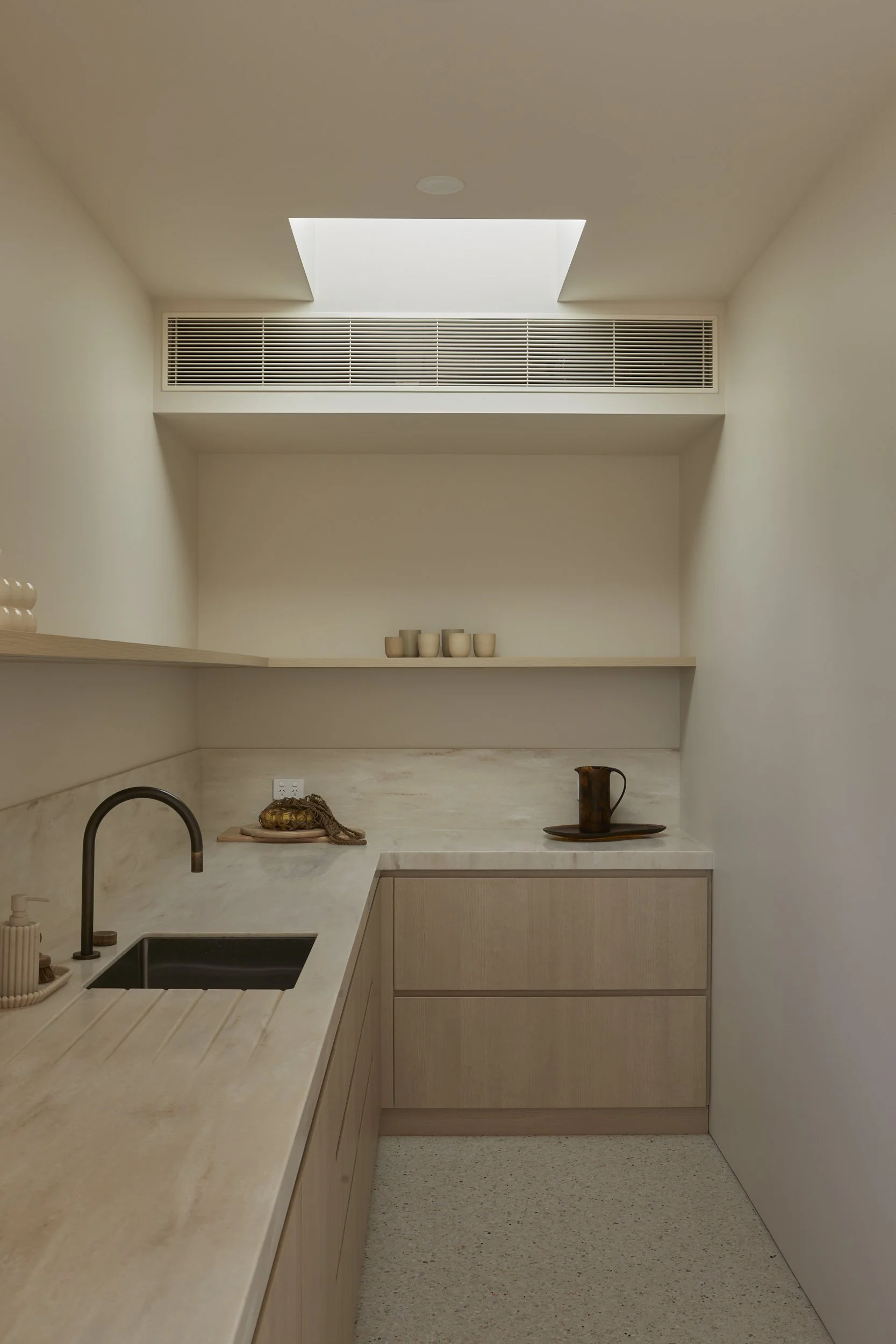 Minimalist kitchen with beige cabinetry, corian benchtops, a sink, open shelf with beige cups, and a skylight providing natural light.