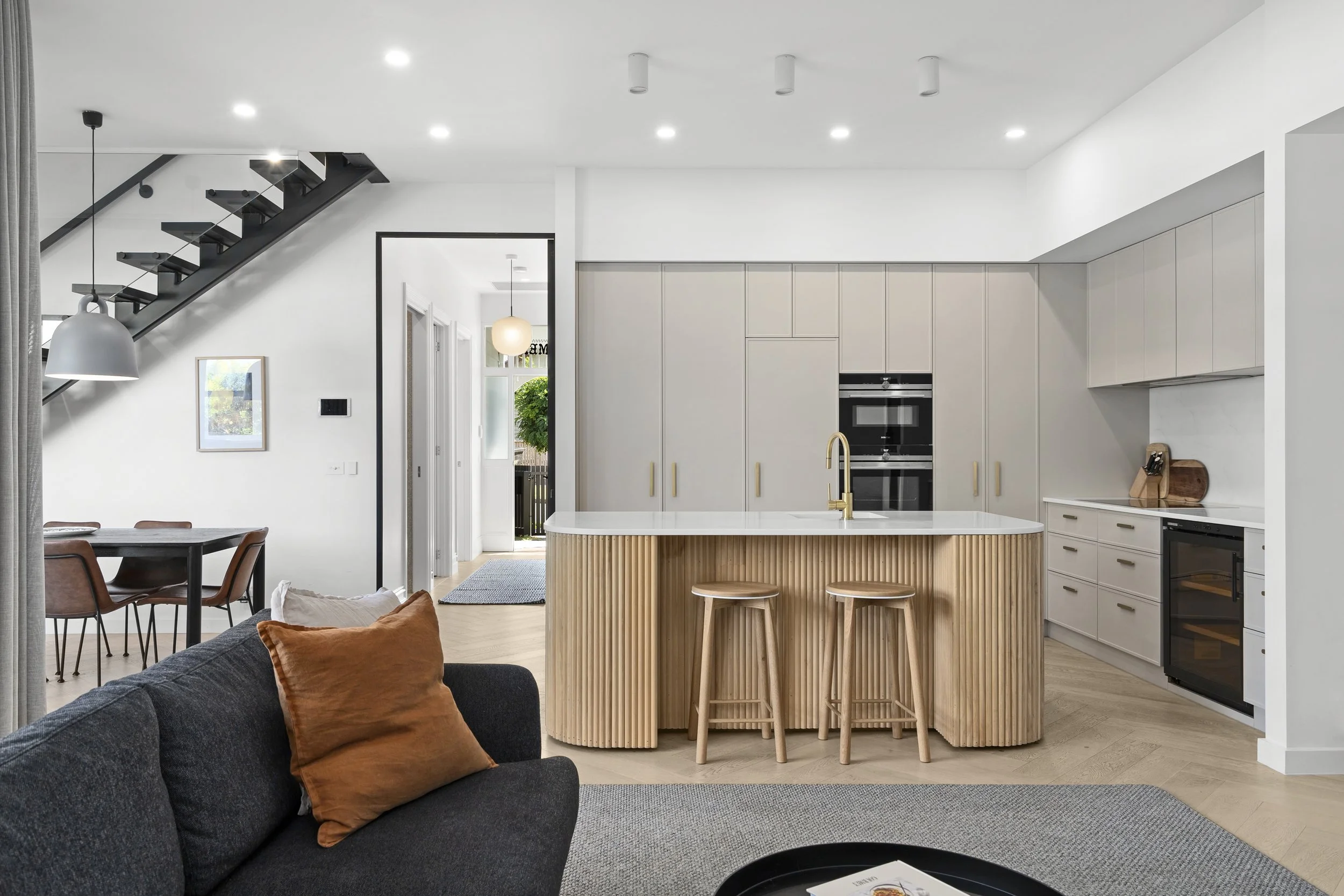 Modern kitchen with white cabinetry, a solid surface staron cotton white kitchen island with a white countertop and wooden paneling, two wooden bar stools, built-in oven, and a black wine fridge. 