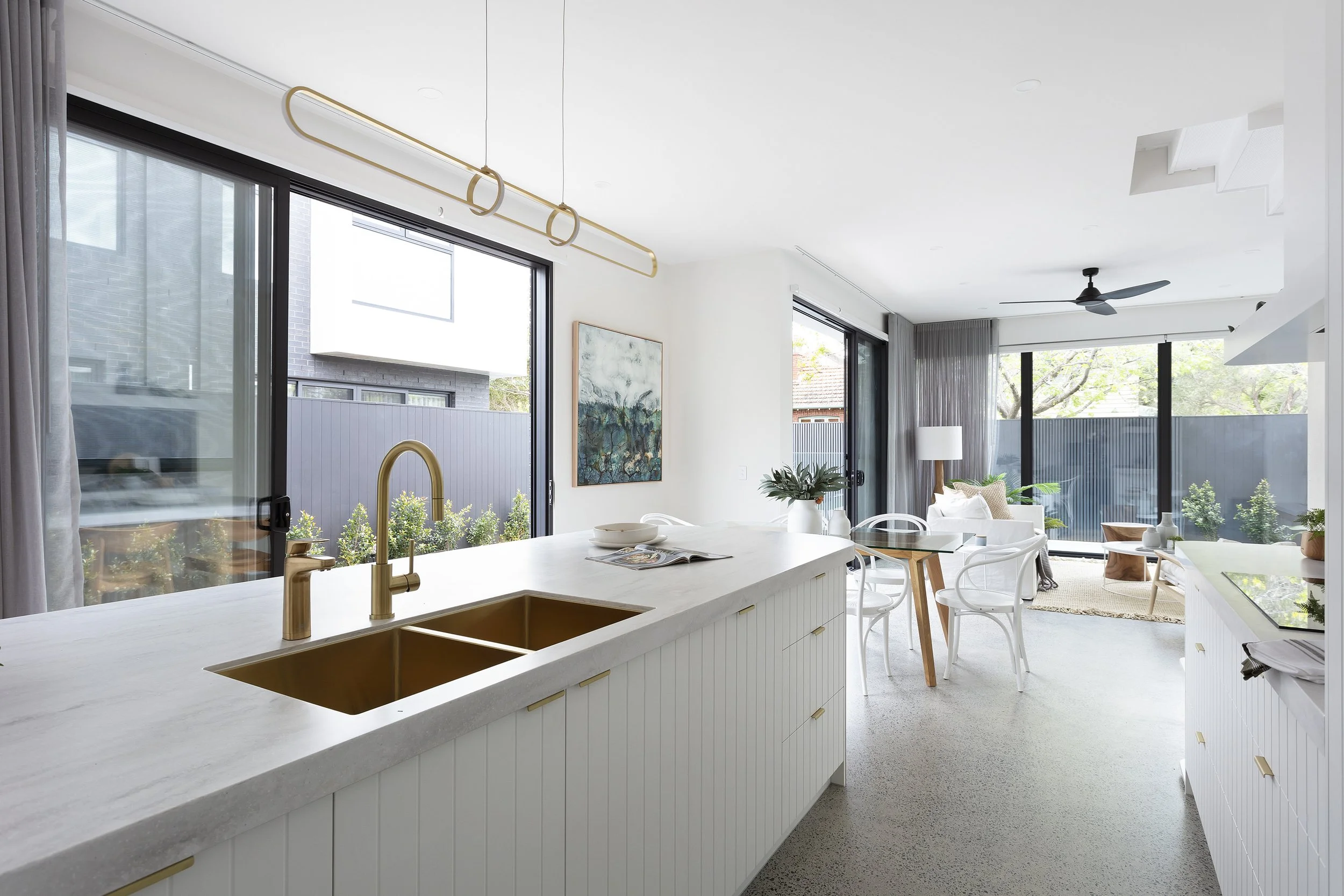 Bright open-concept living space with large sliding glass doors, corian raincloud island with gold sink and fixtures, and a dining area with white chairs and a small table. 
