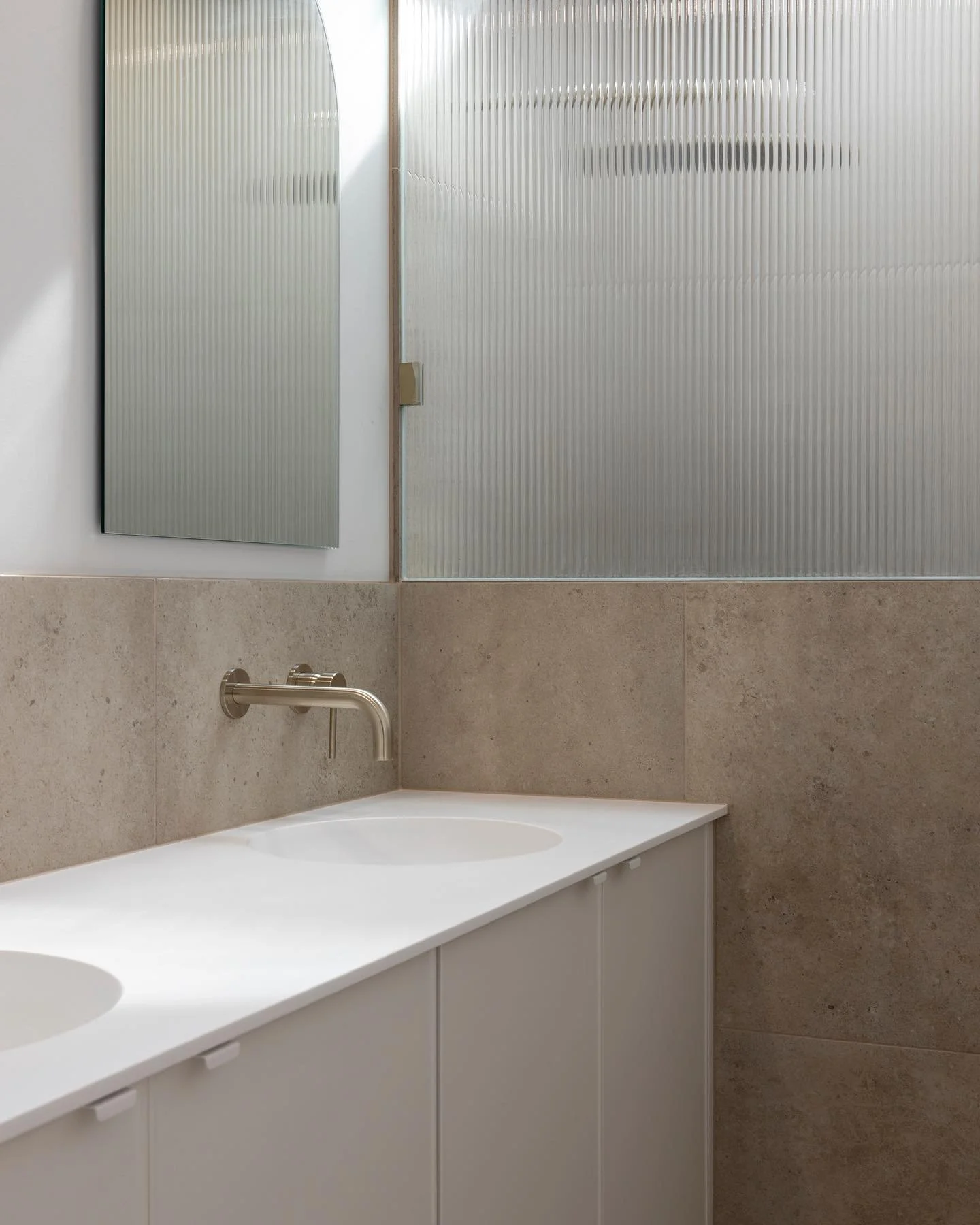 A corian glacier white bathroom vanity benchtop with two integrated basins, and a wall-mounted faucet. Above the vanity, there is a mirror and a textured glass window or partition.