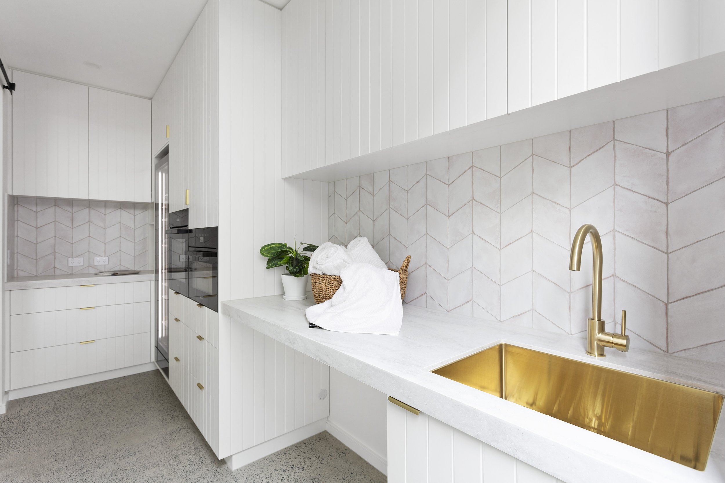 Modern corian raincloud laundry benchtop with brass sink and faucet, plant, and towels on a white countertop with geometric tile backsplash.