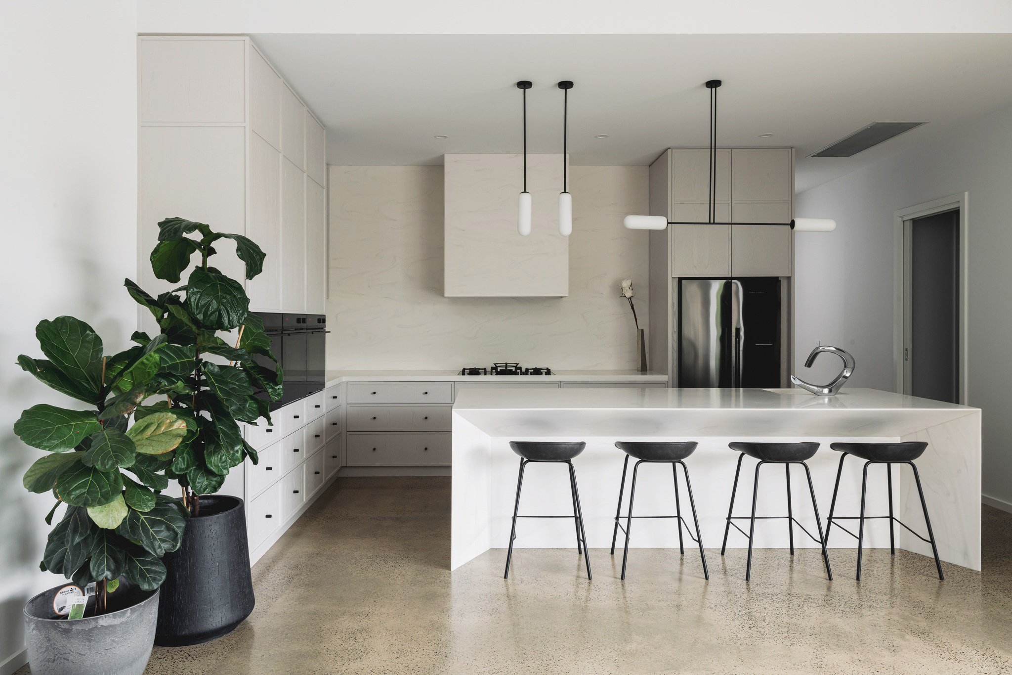 Modern kitchen with white cabinets, a corian valente grigio white kitchen island, black bar stools, stainless steel refrigerator, and a potted plant.