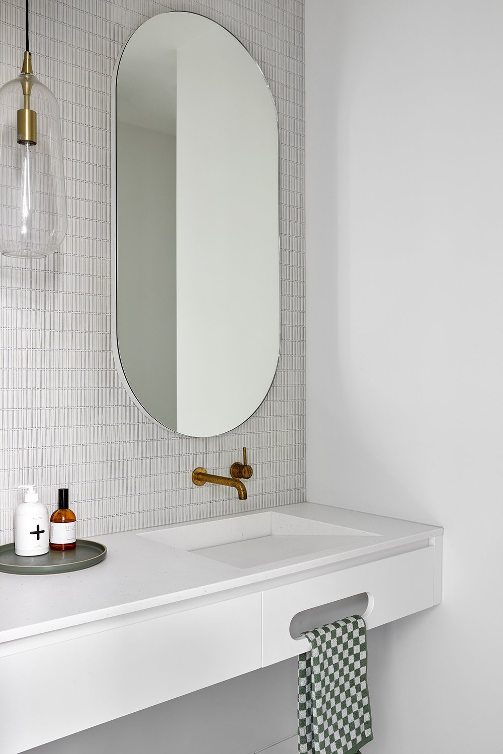 Modern bathroom with a corian glacier white benchtop vanity with integrated basin, oval mirror, gold faucet, hanging glass light fixture, and a green and white checkered towel.