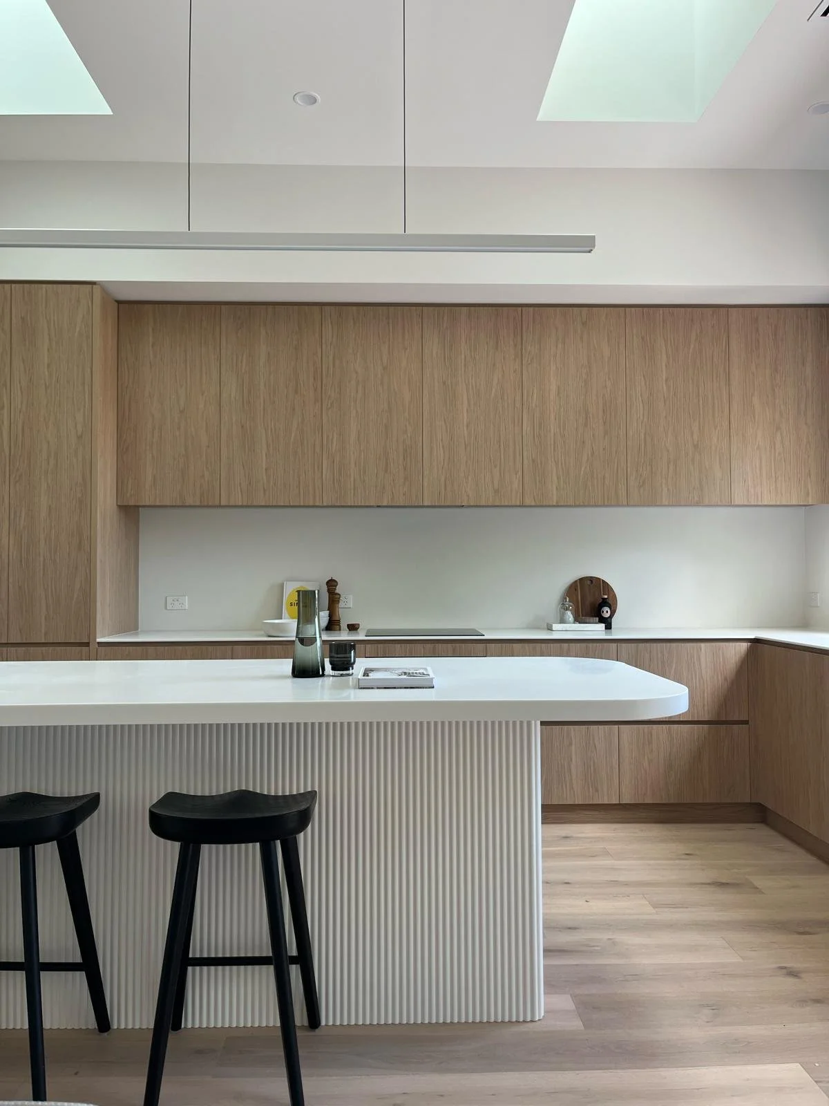Modern kitchen with wooden cabinets, white island with ribbed panel, black bar stools, and skylights in the ceiling.