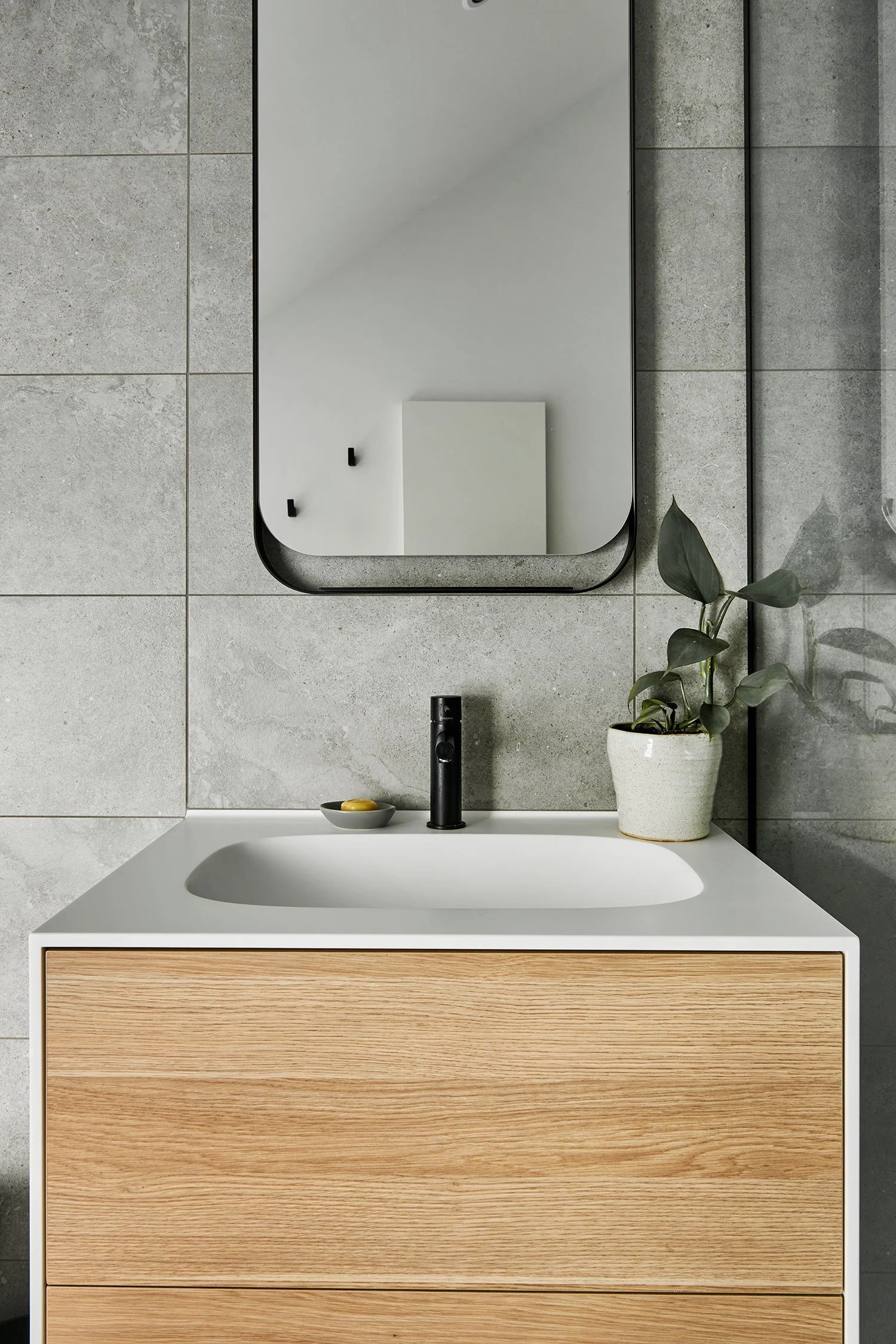 Modern bathroom with a Corian Vanity and integrated basin with black faucet, mirror, gray tiled wall, potted plant, and small dish with soap.