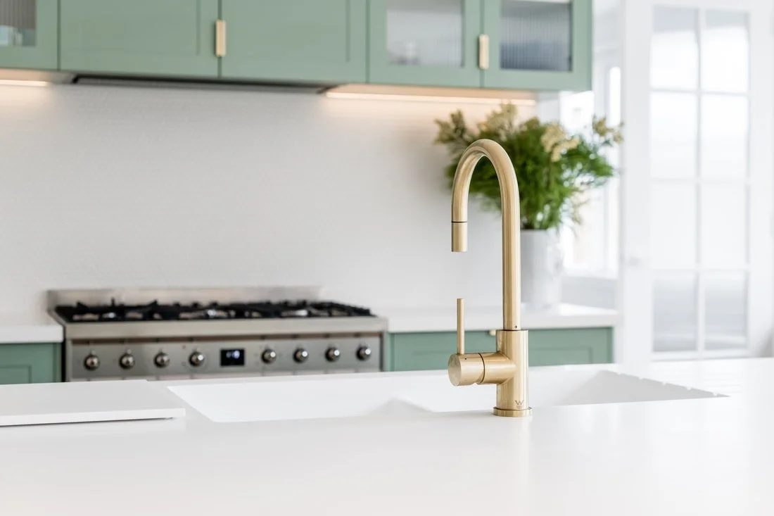 A modern kitchen with a brushed brass faucet and Corian Glacier white benchtop, green cabinets, a gas stove, and a potted plant near a large window.