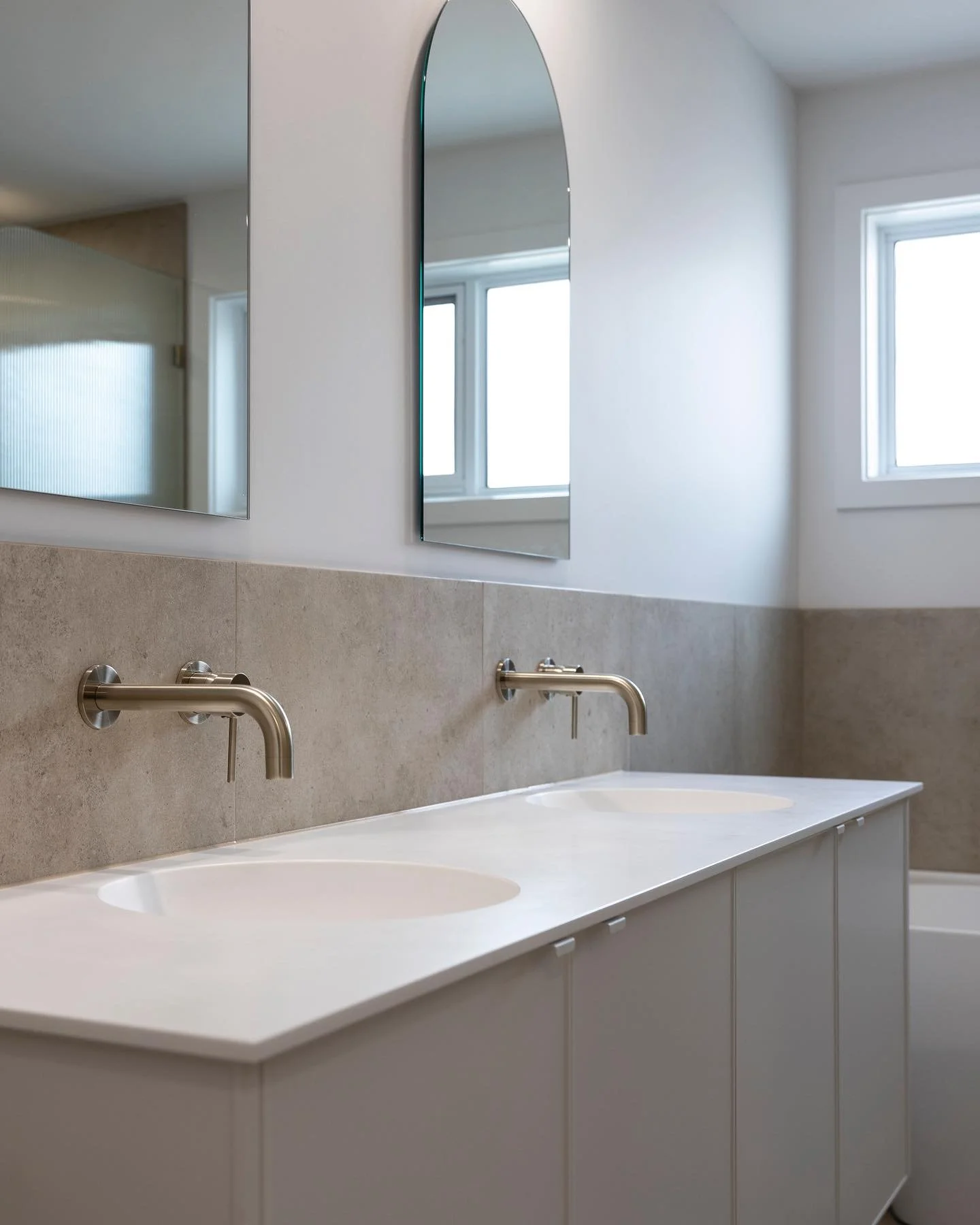 Modern bathroom vanity with dual sinks, wall-mounted faucets, large mirror, and windows providing natural light.