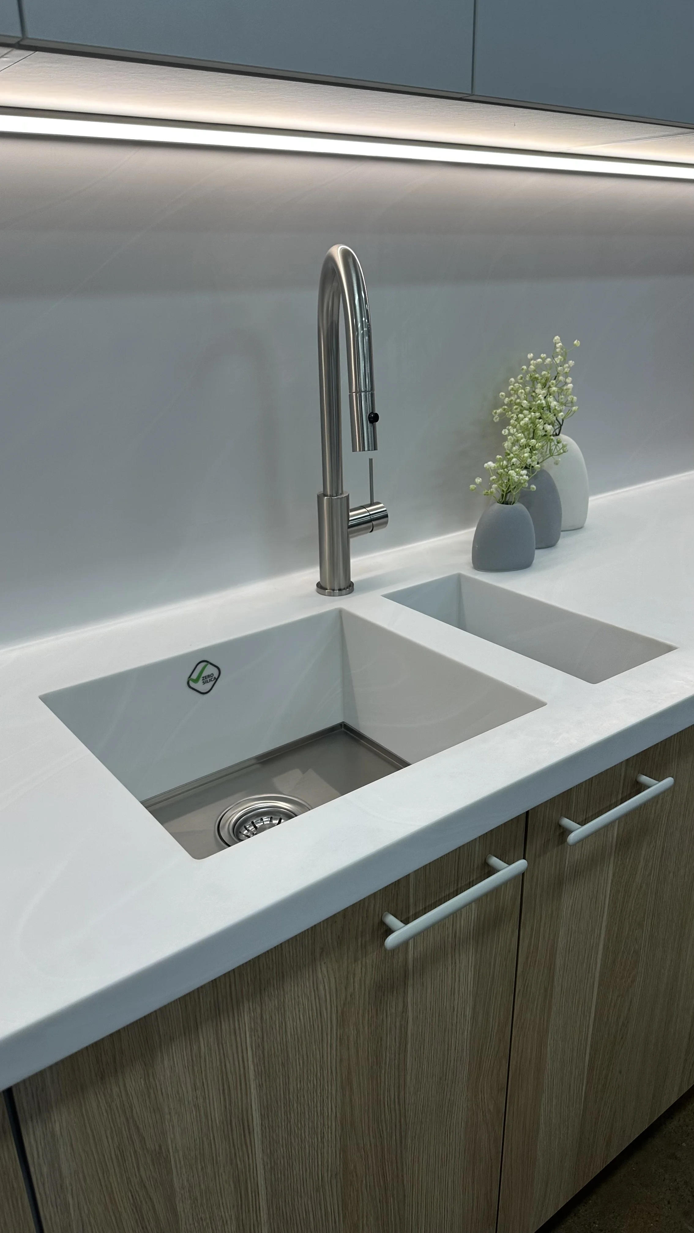 Modern kitchen sink area with double white Corian integrated sink, a stainless steel faucet, and decorative vases with flowers on a white countertop.