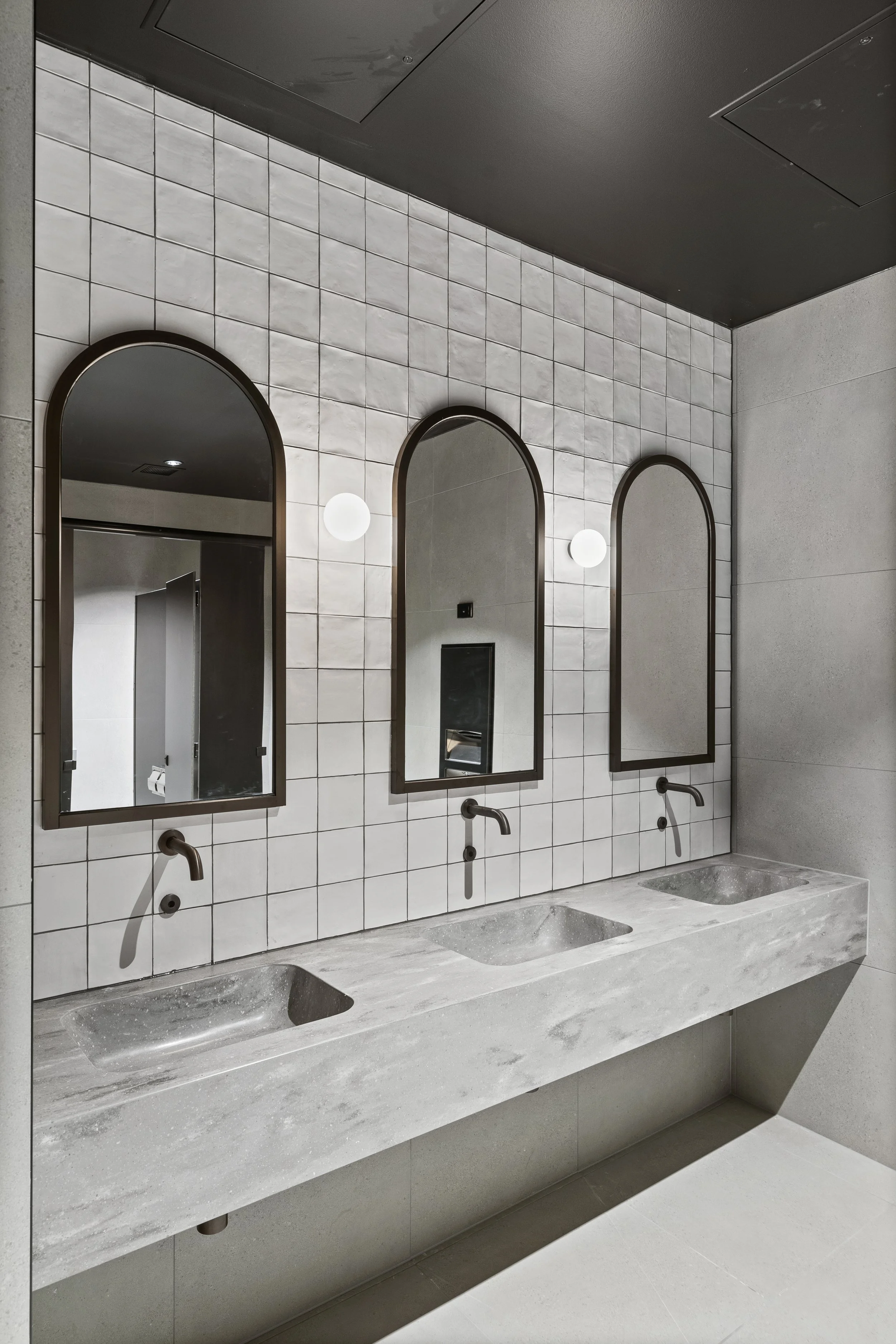 Three mirrors with arched tops and black frames above a grey Corian triple sink with black taps in a modern bathroom with tiled walls and minimal decor.