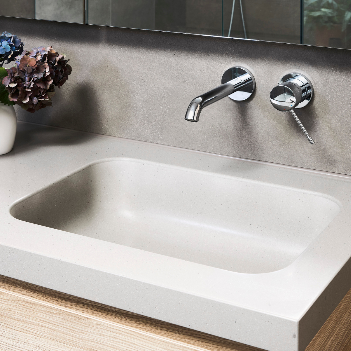 A modern bathroom sink with a chrome faucet and a control handle on a light-colored countertop, with a vase of purple and blue flowers nearby.