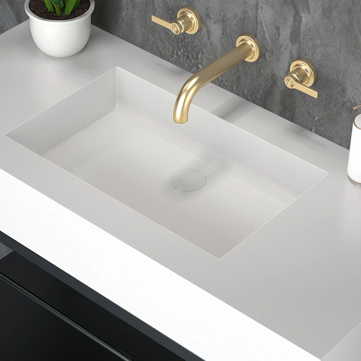 A modern white kitchen sink with a gold faucet and separate gold handles against a gray textured wall, with a potted plant and a soap dispenser on the countertop.