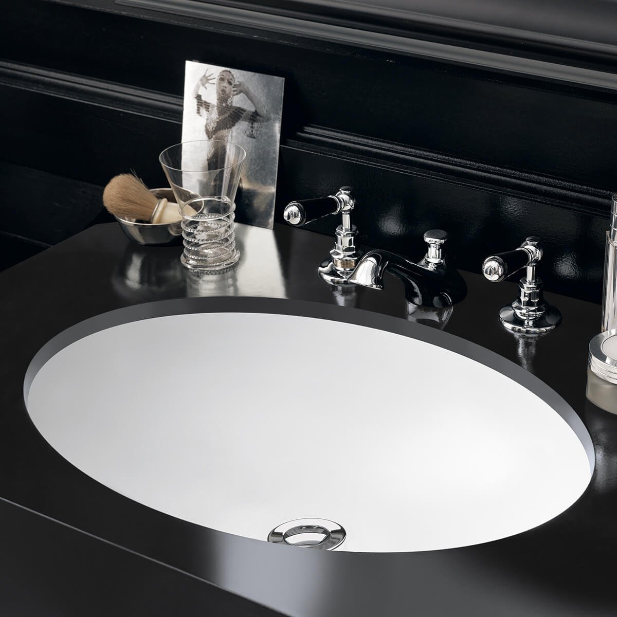 Integrated basin with black Corian benchtop, chrome faucet, and a mirror behind. Items on the counter include a glass, shaving brush, makeup brush, a plastic card with an image of a woman, and a soap dispenser.
