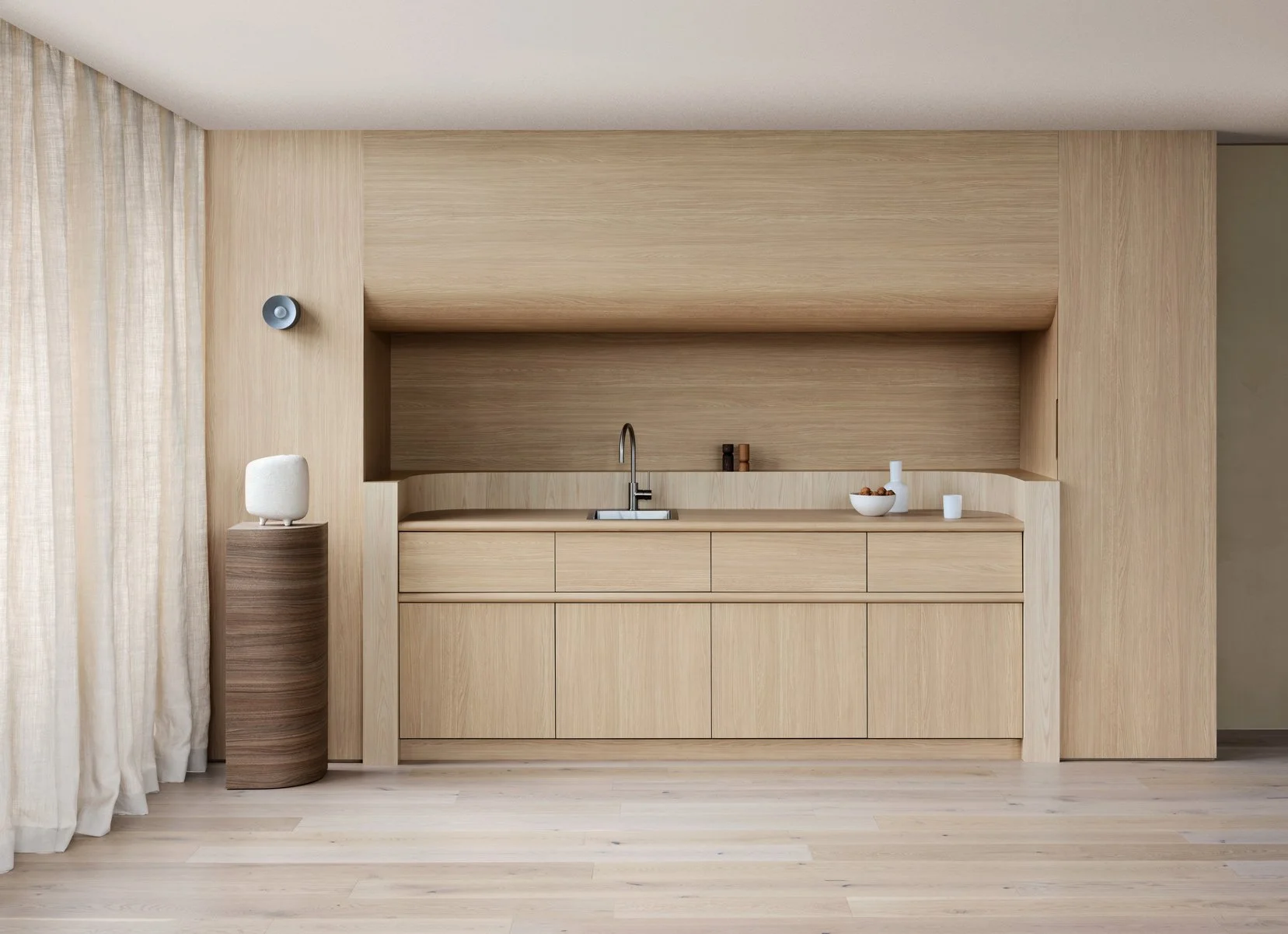 Minimalist kitchen with light wood cabinetry and matching wood walls, a small countertop with a minimalist sink and faucet, decorative white bowls and candles, and a cylindrical stand with a white object on top, located next to a sheer curtain and wi