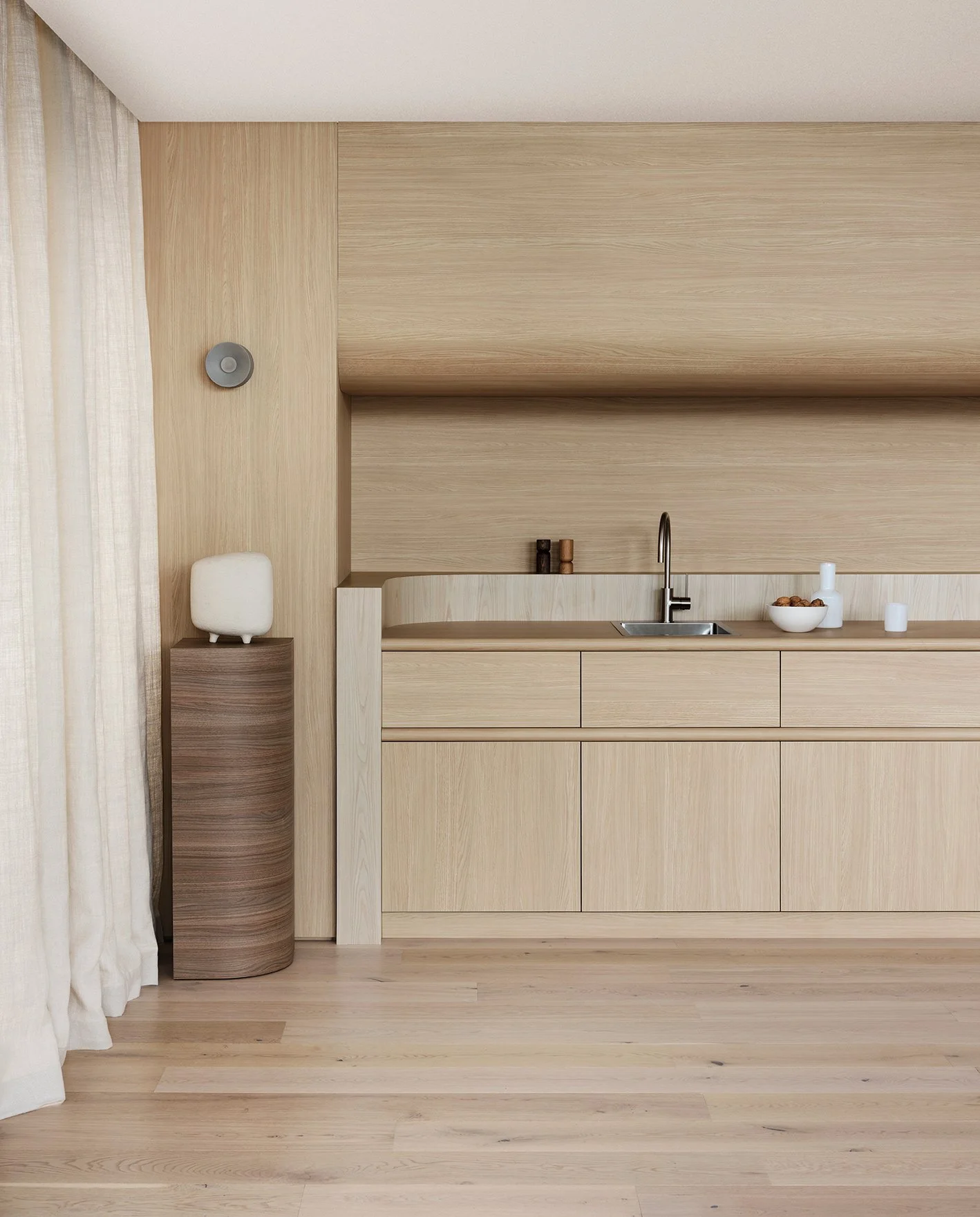 Minimalist kitchen with light wood cabinetry, a small countertop, and a single sink. A bowl of food and small items are on the countertop. A curved wooden pedestal with a white decorative object is to the left, next to cream-colored curtains.